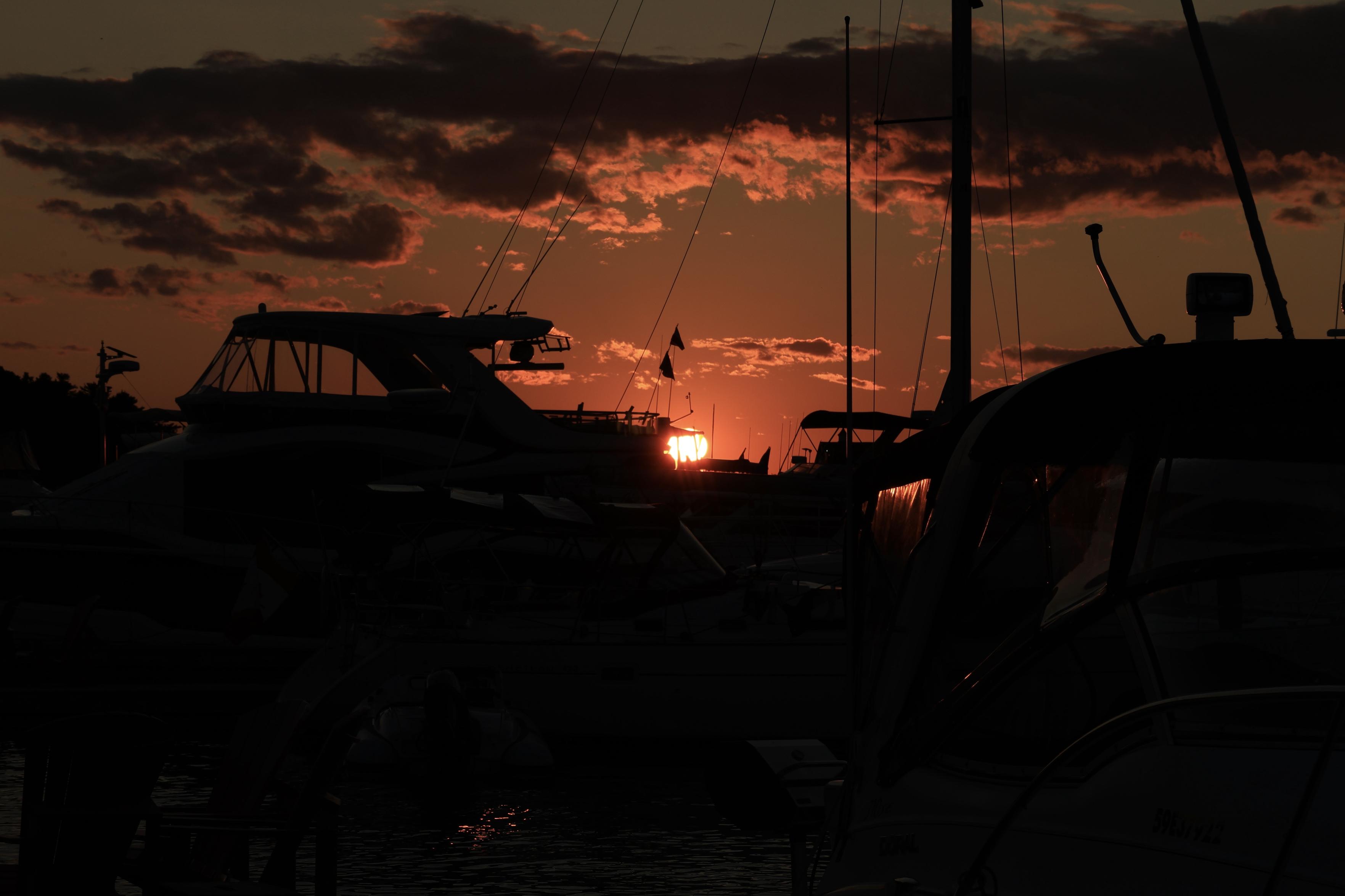 This is a landscape format photo of a sunset taken in the summer from the dock of a marina. The yachts in view are in silhouette with the glowing sun taking centre stage in the photo.  Clouds in the sky overhead are softly illuminated by the warm glow.