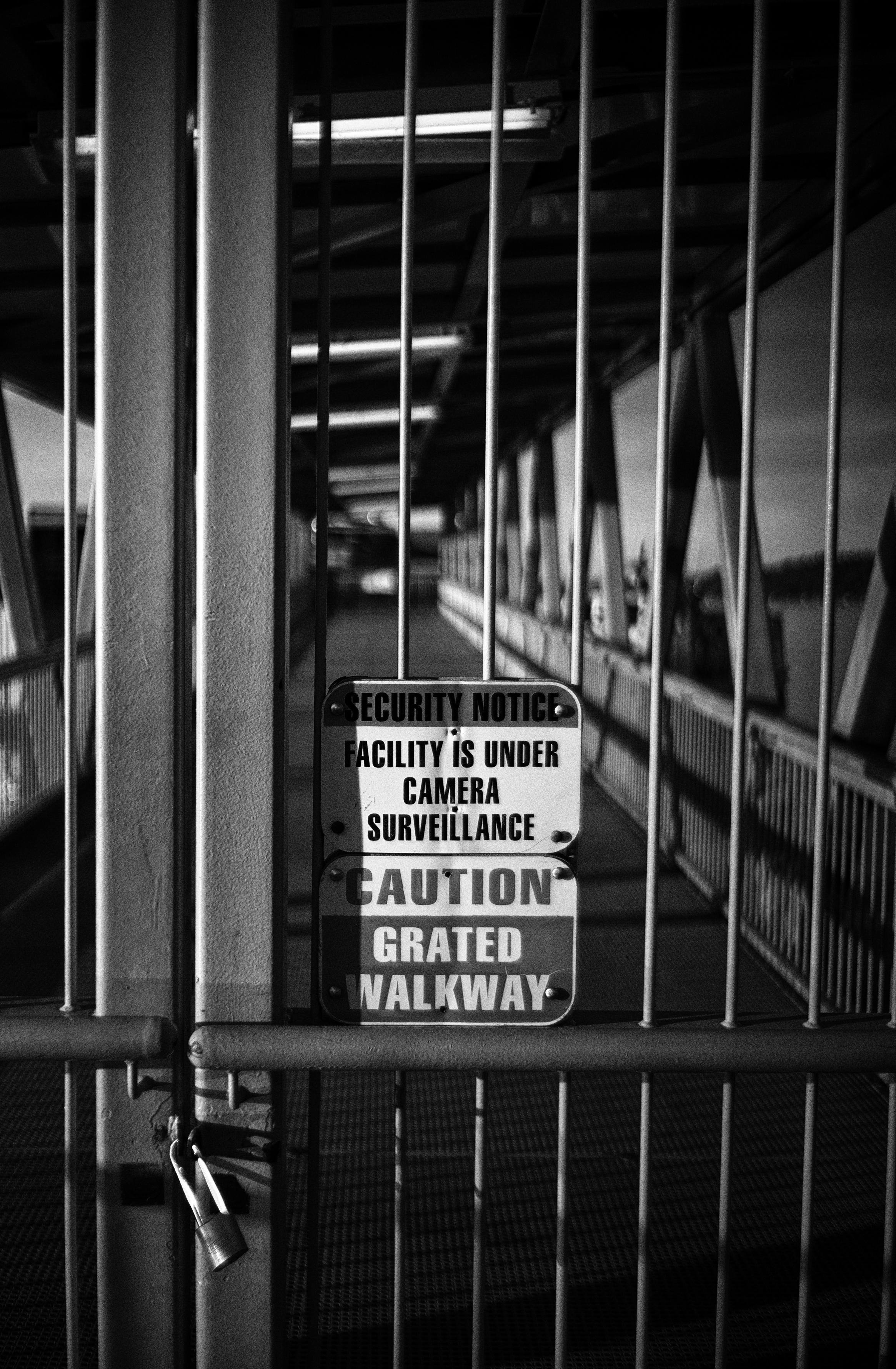 High contrast black and white photo in portrait format of a locked entryway at a ferry terminal.

Two large signs with vertically centered text in the middle of the frame read:

Top sign:
SECURITY NOTICE
FACILITY IS UNDER
CAMERA 
SURVEILLANCE

Bottom sign:
CAUTION
GRATED
WALKWAY

The gate is made of two doors framed with large rectangular beams on its outer sides. The two vertical inner beams of each door are framed in the photo about 20 percent from the left, and are secured by a single padlock about 20 percent from the bottom. A large diameter cylindrical handle running horizontally across the entire width of each door, and small diameter rods run vertically from top to bottom.

In the background the metal grated walkway, side guardrails supported by triangular beams, and roof structure converge into a small rectangular point in the top third of the photo.