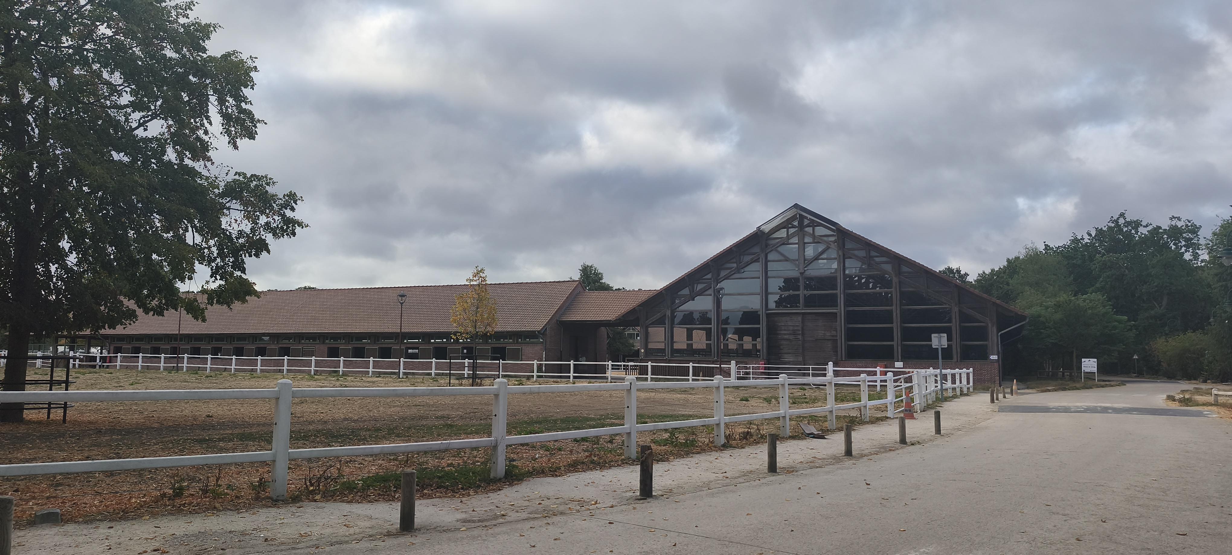 Grand bâtiment d’un centre équestre, avec une haute façade vitrée et une charpente sombre, derrière des barrières blanches. Le ciel est chargé de gros nuages gris et les premiers feuillages d’automne apparaissent autour du bâtiment