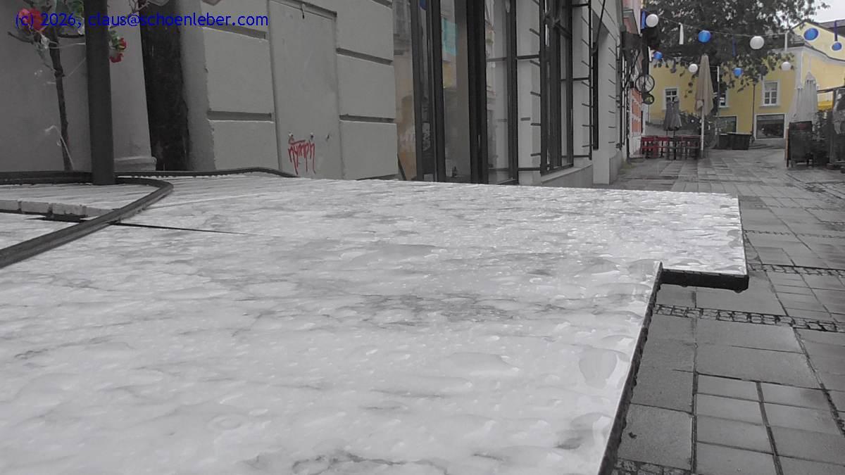 Ein Blick entlang der Gasse vor meinem Büro. Hier sind normalerweise ein halbes Dutzend Cafés in Betrieb. Aber seit gestern regnet es, und niemand will heute auf der Straße sitzen und Kaffee trinken oder zu Mittag essen.

Im Vordergrund des BIldes stehen vier Tische, zusammengerückt, weil das Café geschlossen ist. Auf den marmorartigen, weißen Flächen der Tische steht Regenwasser in kleinen Pfützen. Im Hintergrund erstreckt sich die Gasse und zeigt im weitere Verlauf zwei ebenfalls geschlossene Cafés, deren Tische auch zusammengerückt darauf warten, dass der Regen aufhört.

Die Gasse ist in Passau (Niederbayern) und heißt Grabengasse.