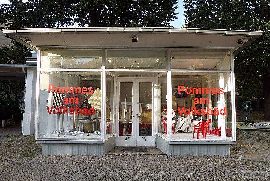 Ein kleiner, freistehender weißer Pavillon mit Schaufenstern und in der Mitte ein Eingang auf dem Rosa-Luxemburg-Platz neben der Volksbühne. Auf den Scheiben der Schaufenster rechts und links die Aufschrift „Pommes am Volksbad“ in roten Buchstaben in einer fetten Grotesk-Schrift.