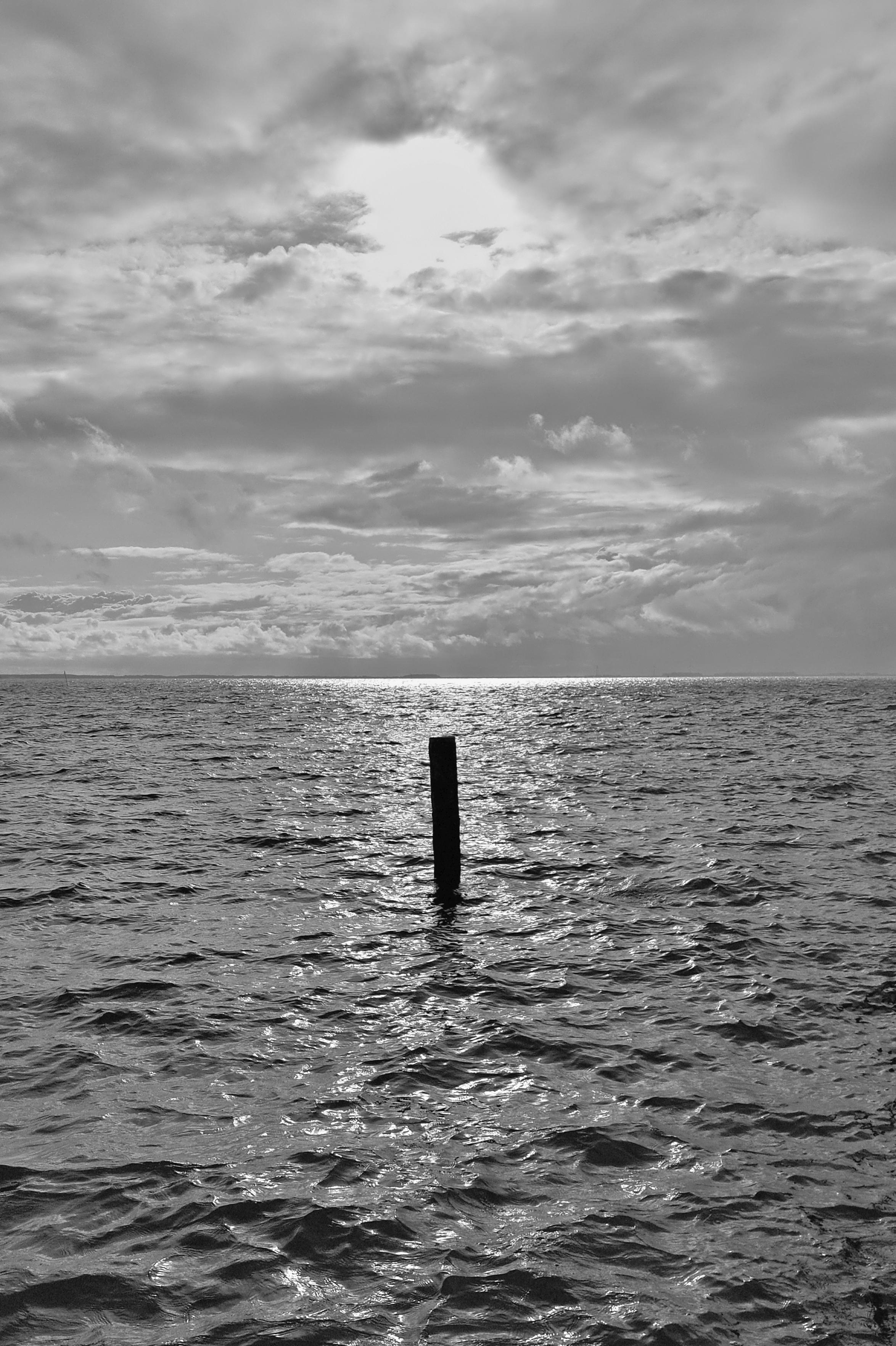Schwarzweiss-Foto des Wattenmeeres bei Flut als Teil der Nordsee. Sonne scheint als Gegenlicht von vorne durch die Wolken. In der Mitte des Bildes ist ein Festmacher-Dalben in Schwarz zu sehen.