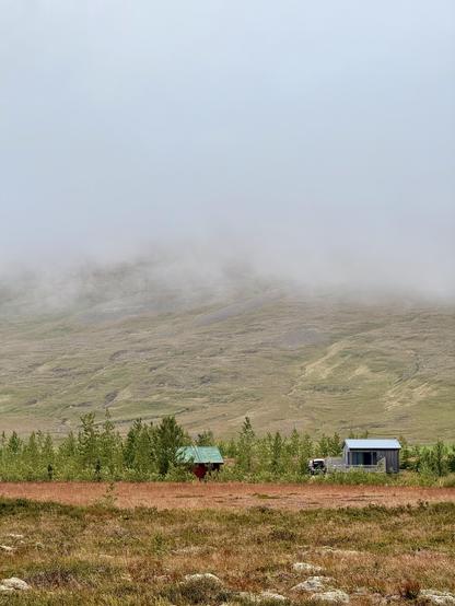 Two small cabins, one with a green roof and one gray, sit in front of a line of trees and a mountain partially covered in fog.