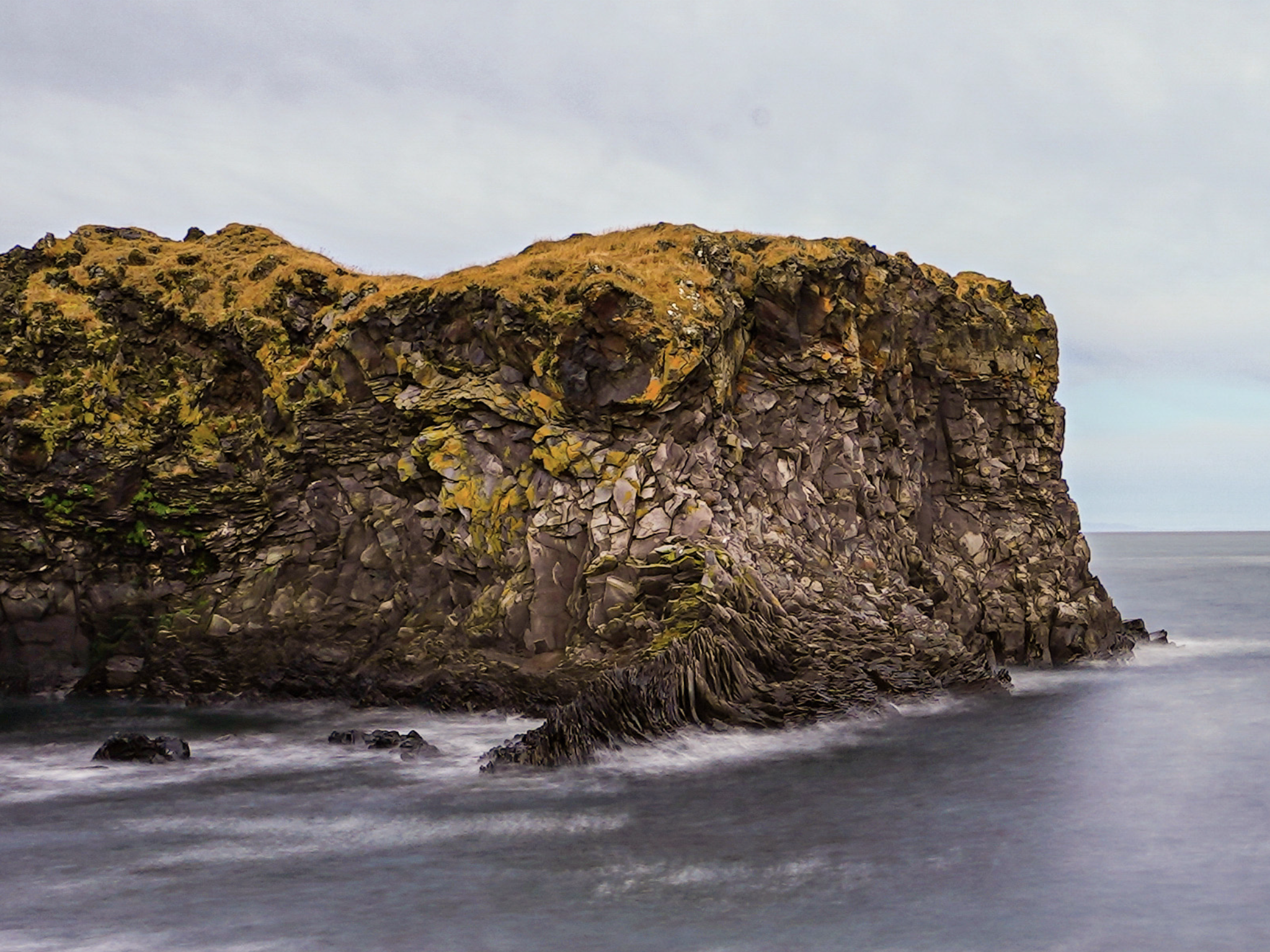 Large rocky land part in the sea. Yellowish moss on top, some green spots, brown greyish rocks. The blueish sea is fuzzy, due to a long shuttertime.