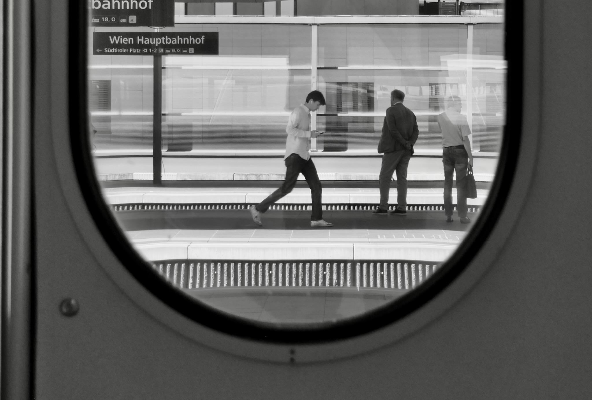 Photo of a walking young man, white shirt, black trousers, sneakers. He is walking down the platform in the Vienna Main Station, holding a mobile in front of him. Next to him two men are waiting for a train, back turned to the point of view.
Picture was taken through a ICE train window, which is round on the bottom. Photo is black and white.