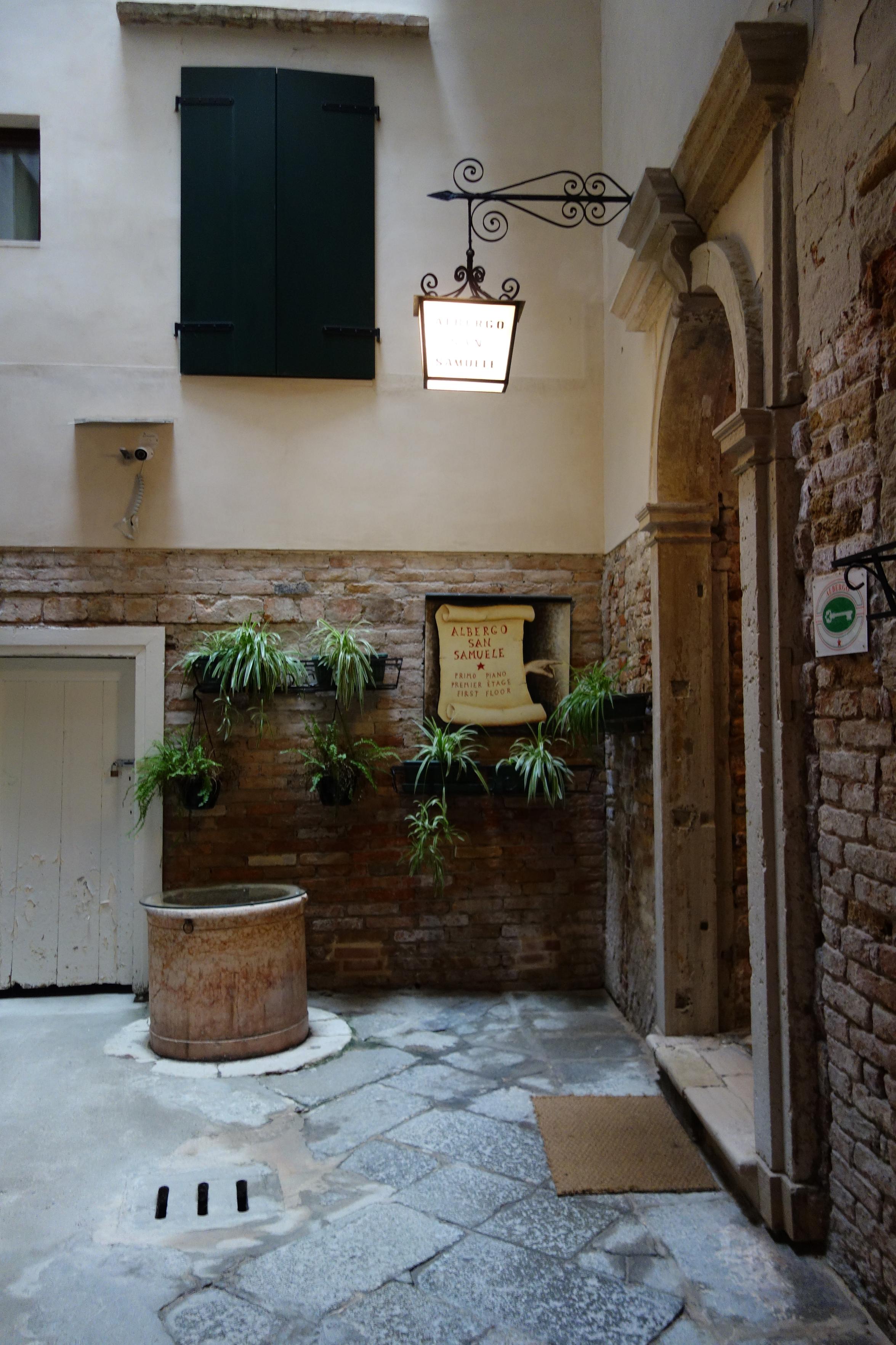 A hall in Venice with a small iron lamp above the door with some plants on the wall and a text as an old document next to it.