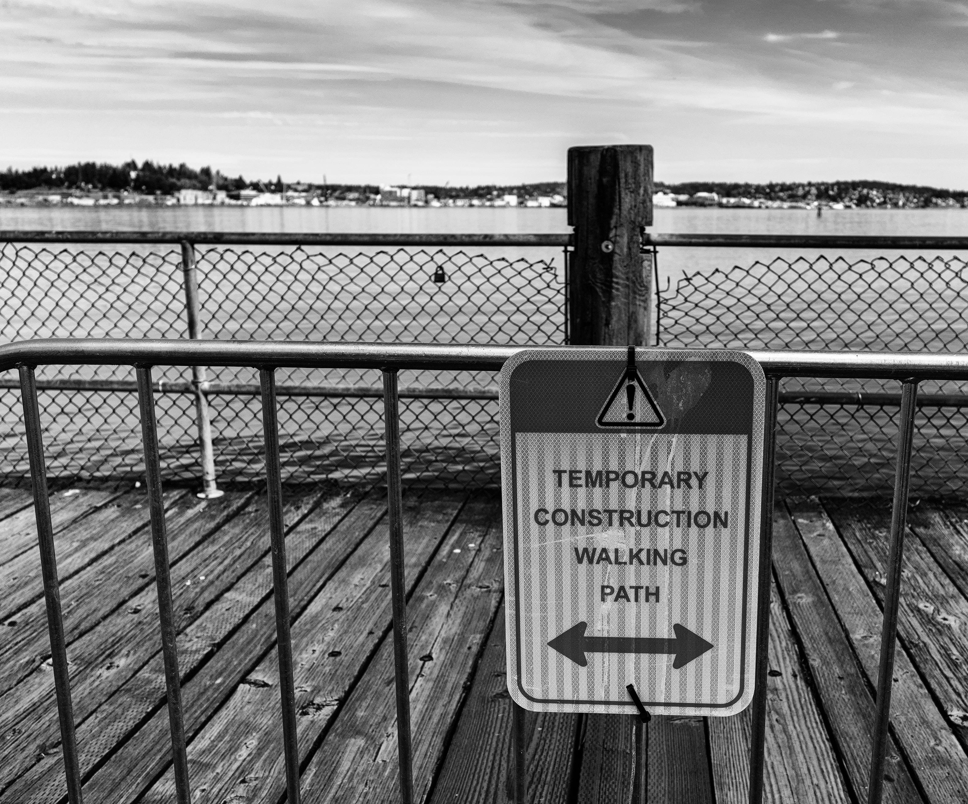 Black and white photo in landscape format of a closed & barricaded walking path closed due to safety concerns.

A temporary steel mobile barricade stretches horizontally across the bottom half of the photo with 8 evenly spaced vertical support bars. A warning sign is attached to the 4th to 2nd support bars counting from right to left that reads:

[black and yellow triangular warning icon with exclamation point]
TEMPORARY
CONTRCUTION
WALKING
PATH
[large double sided arrows]

Behind the barricade are dark colored wooden planks perpendicular to the bottom   of the photo and a chain link safety rail fronting the ocean. An ocean inlet and coastline are in the far background with cirrostratus clouds looming above.