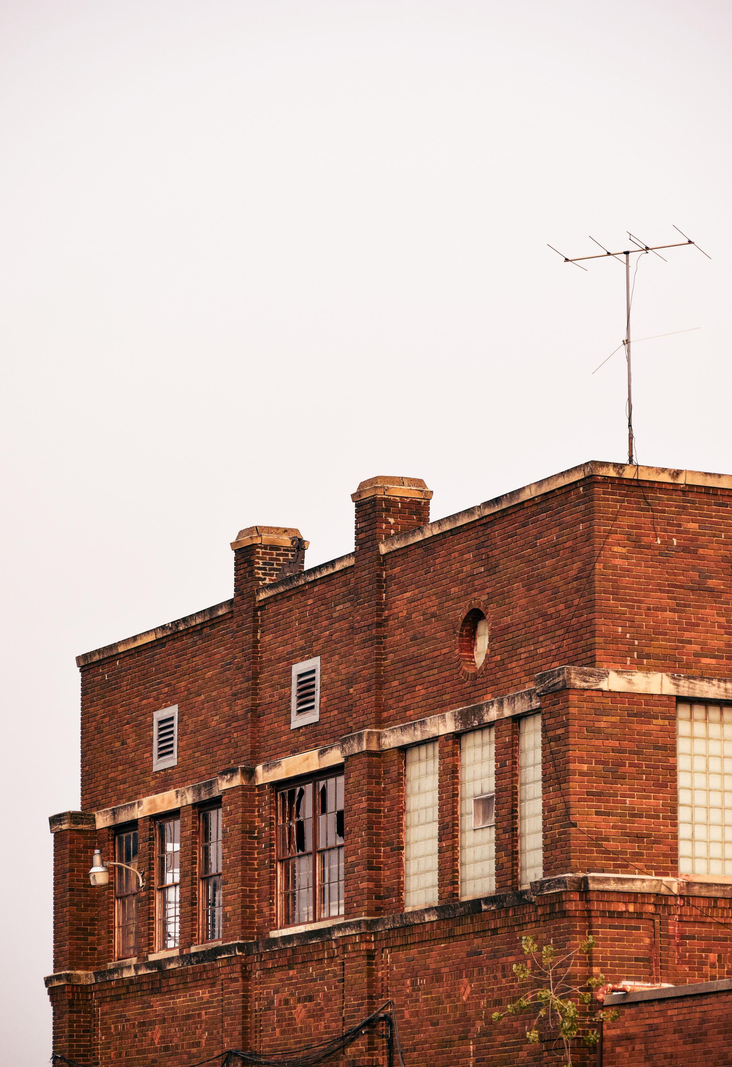 Weathered red-brick industrial building with tall windows, chimneys, rooftop antenna, and autumn foliage against a pale sky; quiet, historic mood.