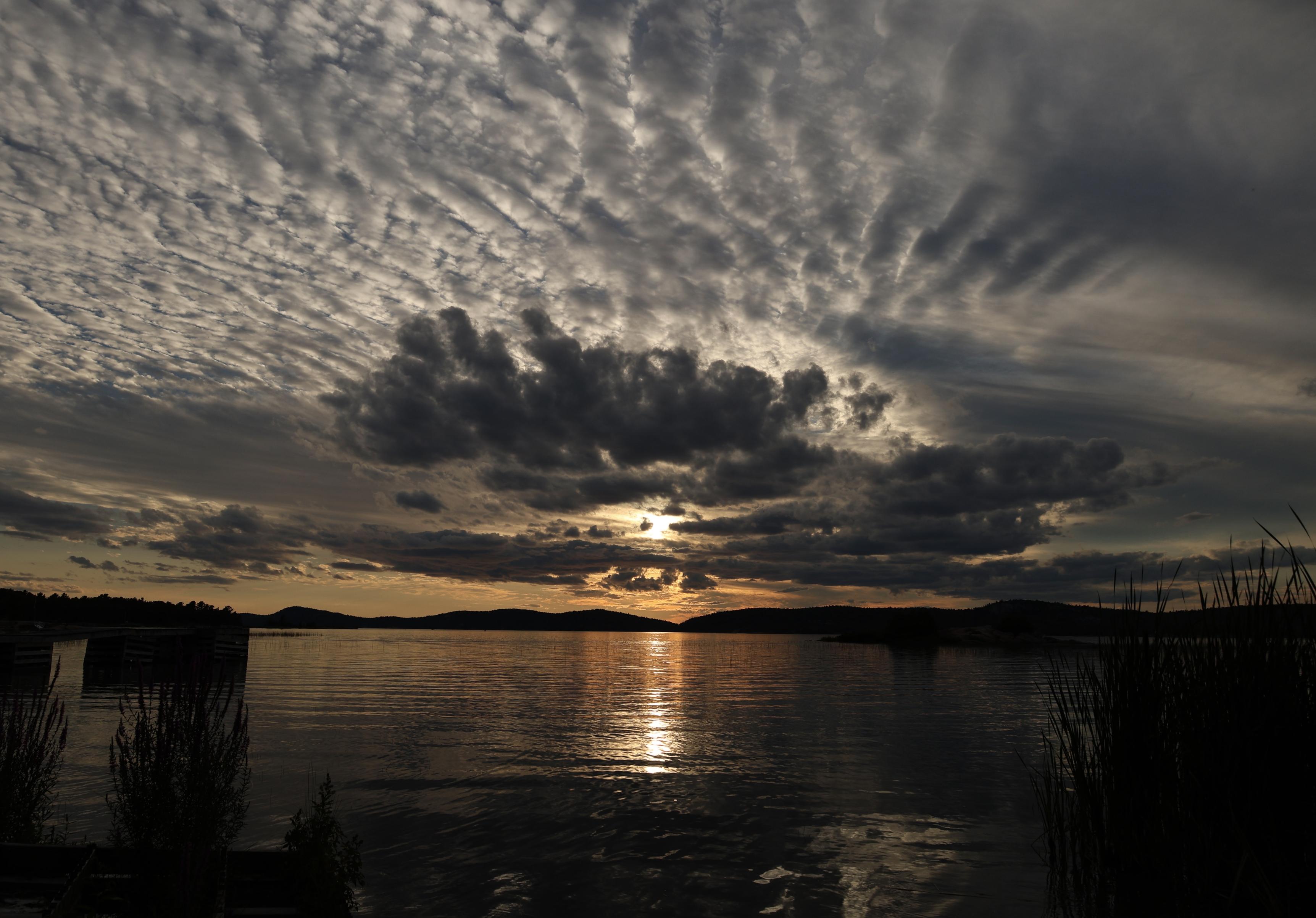 This is a landscape format photo taken on a summer evening of a sunset over the bay of a larger lake. The water is quite calm and mostly flat, with ripples visible in the reflecting sunlight. Low range mountains in the distance can be seen just above the water's horizon line in the photo. The sun is surrounded by clouds but still glows in the centre of the photo. The clouds overhead are a mix of illuminated colour and darker shades. The photo definitely has a warm glow to it.