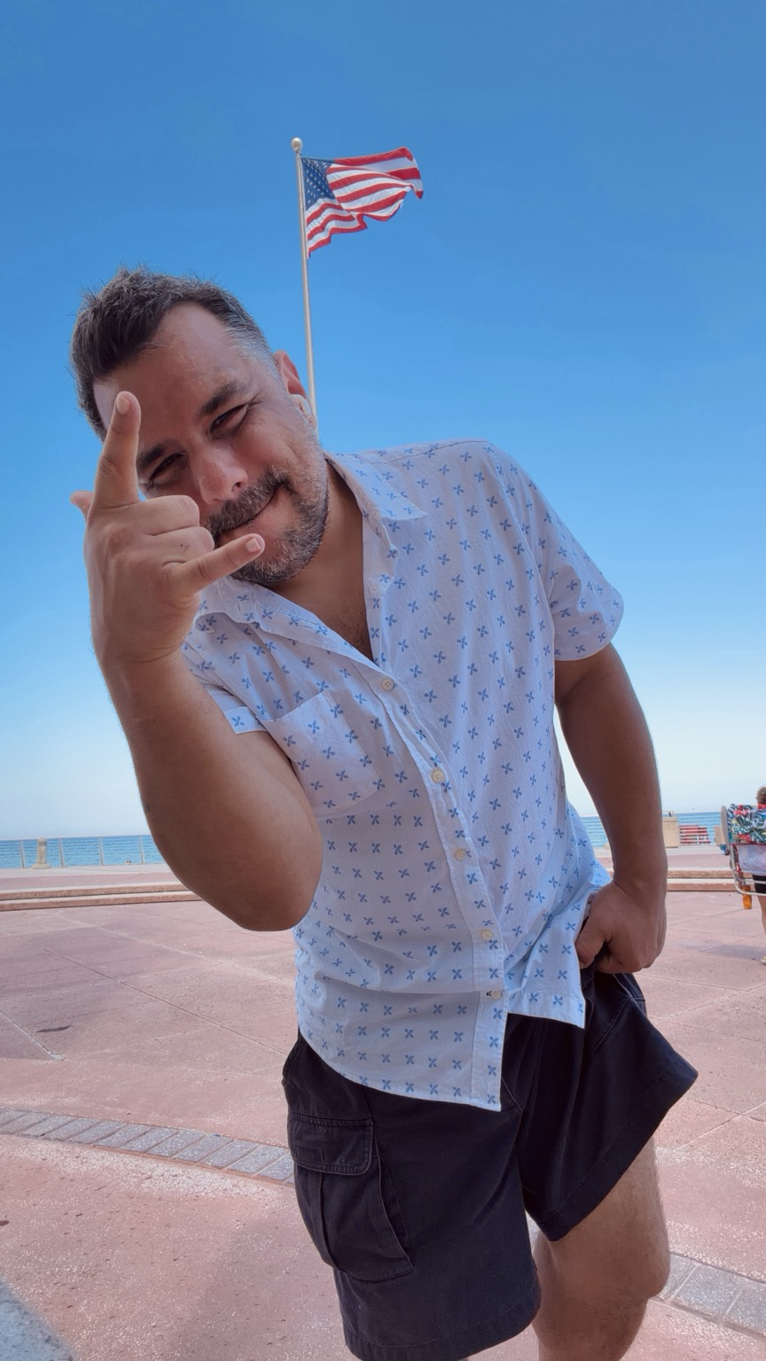 Me wearing a white button up with blue pattern outside, blue sky American flag waving behind me above me