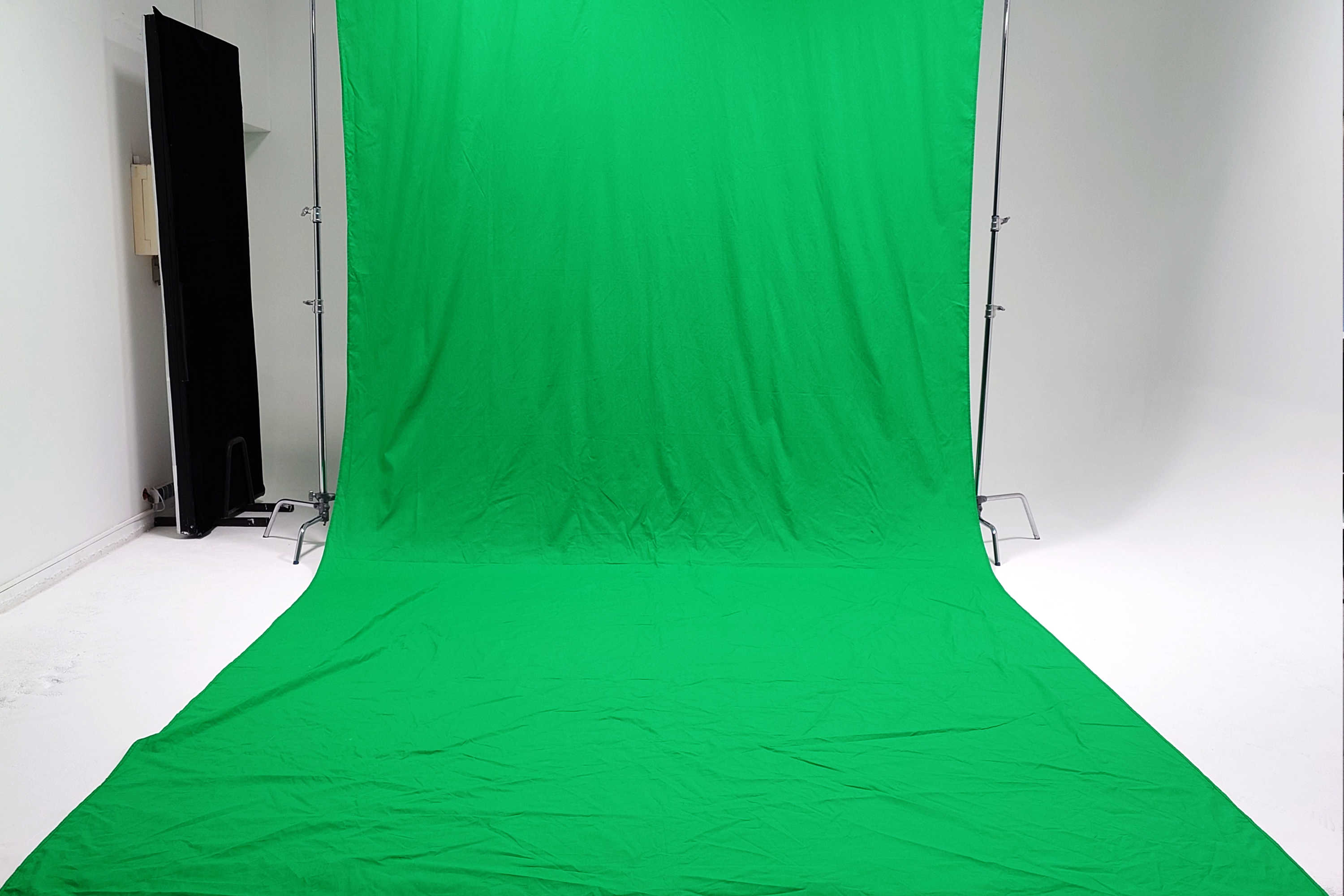 Fotografia przedstawia gotową do użytku instalację green screenu w jasnym studio fotograficznyn i filmowym - Studio Stawki. Na środku, rozpięta na dwóch wysokich metalowych statywach (C-standach), wisi wielka, zielona płachta materiału. Tkanina jest lekko pomarszczona, a jej dolna część spływa na białą podłogę studia, tworząc cykloramę. Po lewej stronie widać czarny, prostokątny element, prawdopodobnie flaga fotograficzna. Tło stanowi biała ściana studia. Całość jest dobrze oświetlona.