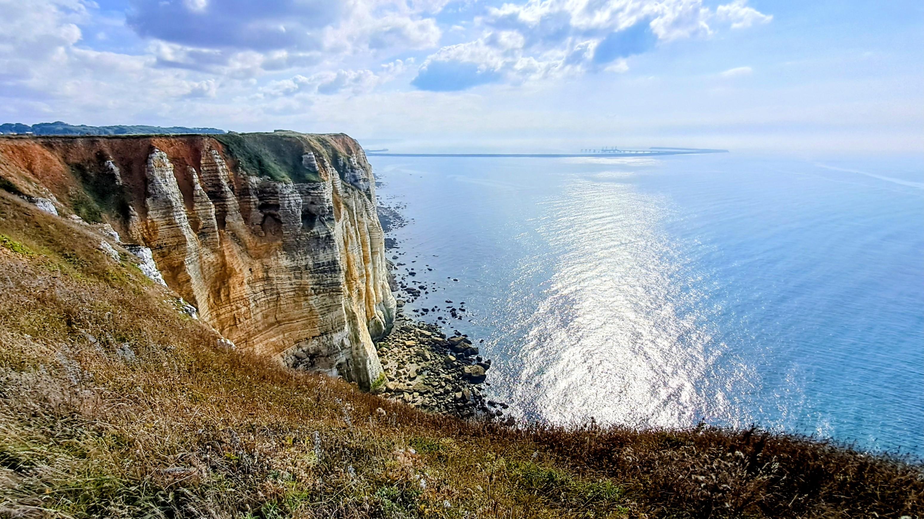 Blick von der Steilküste aufs Meer. Nach links blickt man auf den steilen Abhang mit unterschiedlich farbigen Schichten. Im Vordergrund ist Gras zu sehen. Das Sonnenlicht reflektiert in der relativ glatten Oberfläche des Meeres. Am Himmel sind einige, kleine Wolken zu sehen.