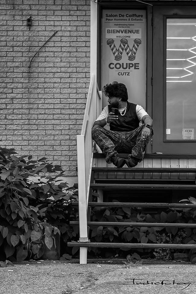 A bearded man with tousled hair is waiting outside a barber shop.