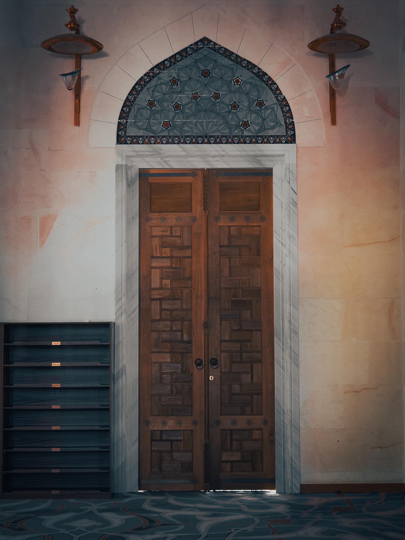 An ornate wooden panelled door, with a beautiful mosaic above with an funky candelabra on either side. Next to the door on the left sits an empty storage unit in dark wood. The photograph was taken at the Grand Çamlıca Mosque (Büyük Çamlıca Camii) in Istanbul, Türkiye.