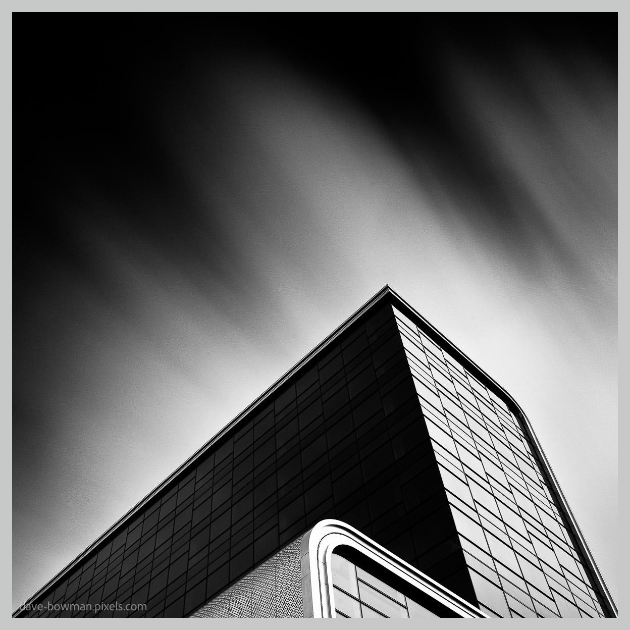 A black and white, long exposure photograph of the RAI Theatre in Amsterdam, showing a dramatic section of the modern building rising sharply into the sky. Dark glass panels and strong angular lines form bold geometric shapes, while the solid, structured architecture contrasts with the softer, slightly changing sky behind it.