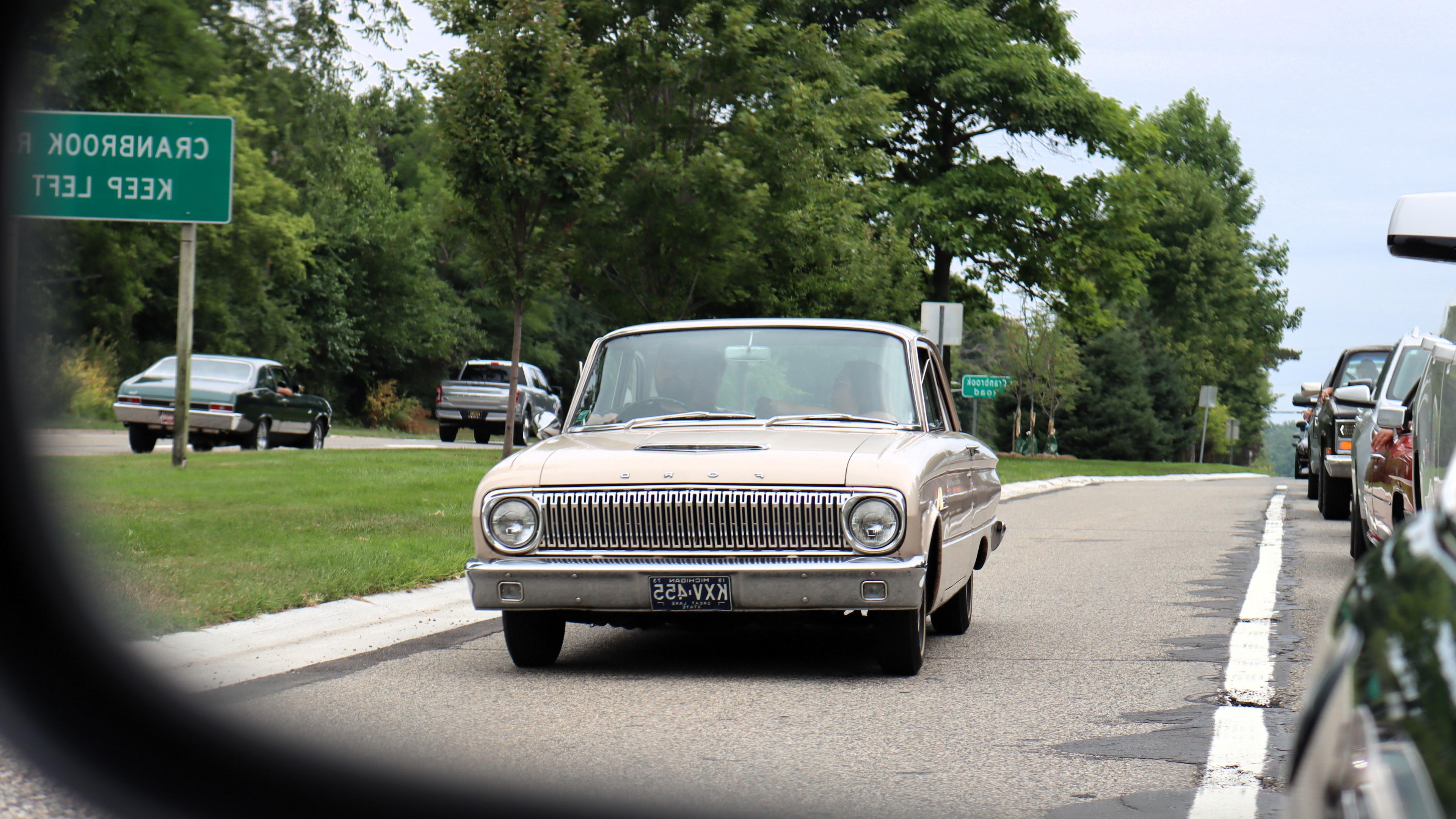 Photograph of an early 1960s Ford Falcon coming from behind in a clear traffic lane as seen in the left side mirror during the 2026 Dream Cruise.