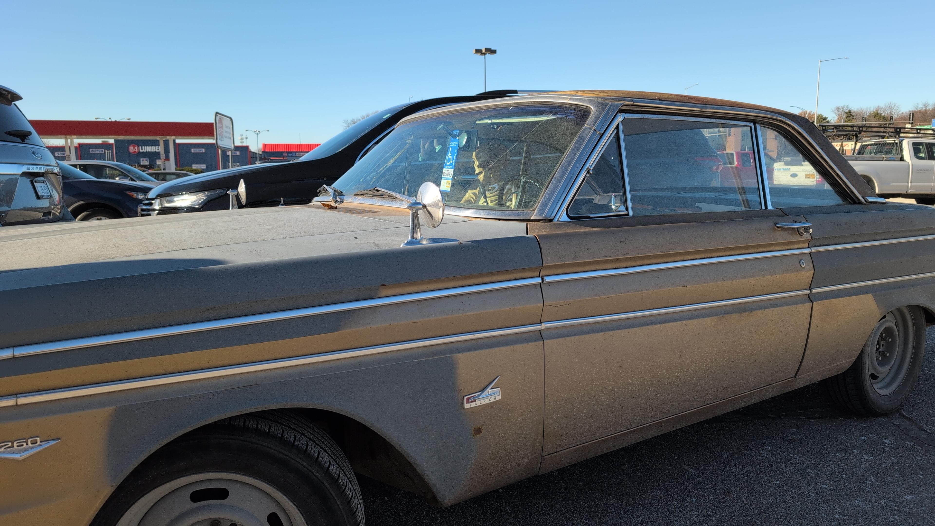 A beige second-gen Ford Falcon sits in a parking lot. A handicapped parking tag dangles from the rearview mirror and a life sized human skeleton model is sitting in the passenger seat.