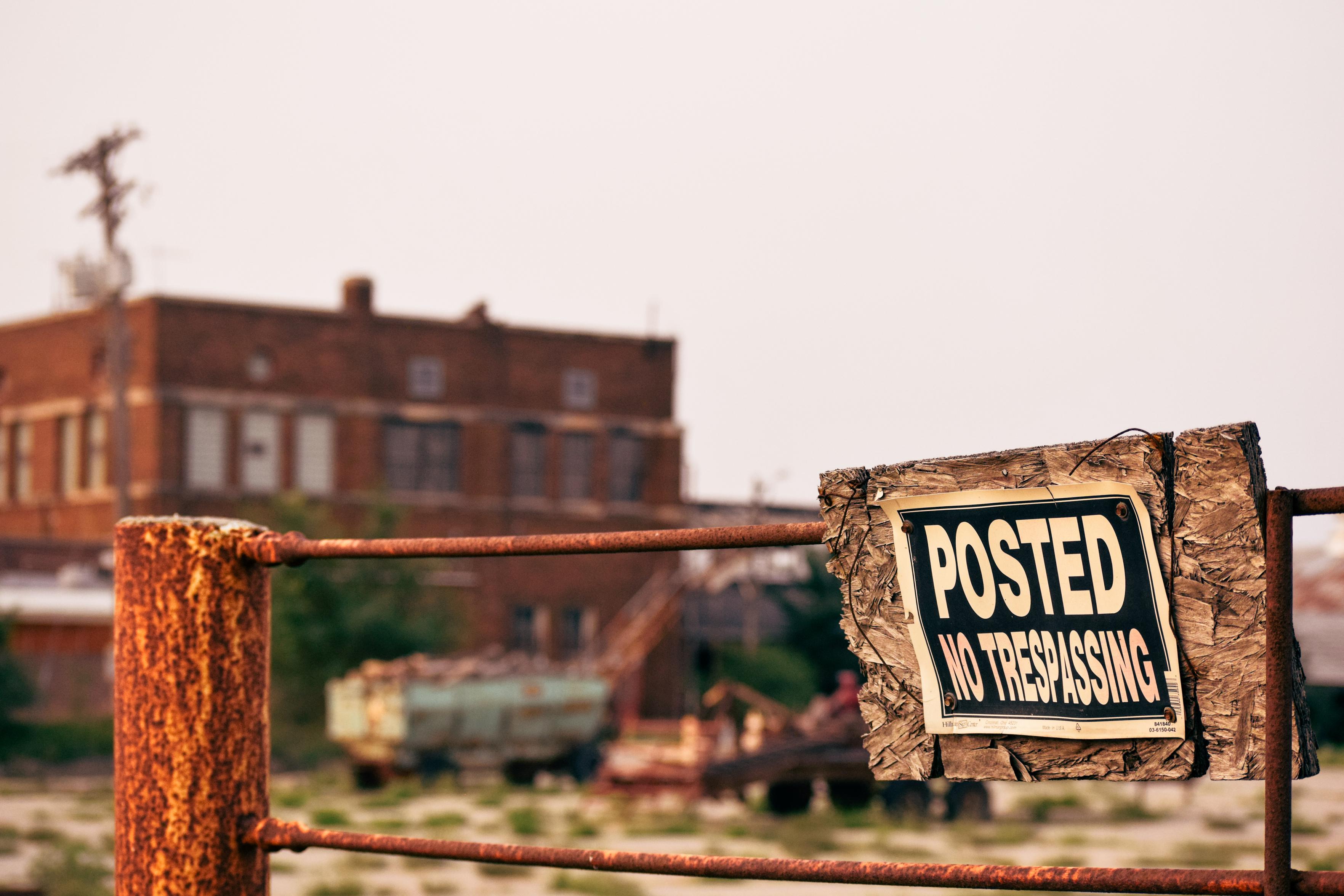 Rusty fence with a weathered “Posted No Trespassing” sign in front of an abandoned brick building, evoking an isolated, cautionary mood.