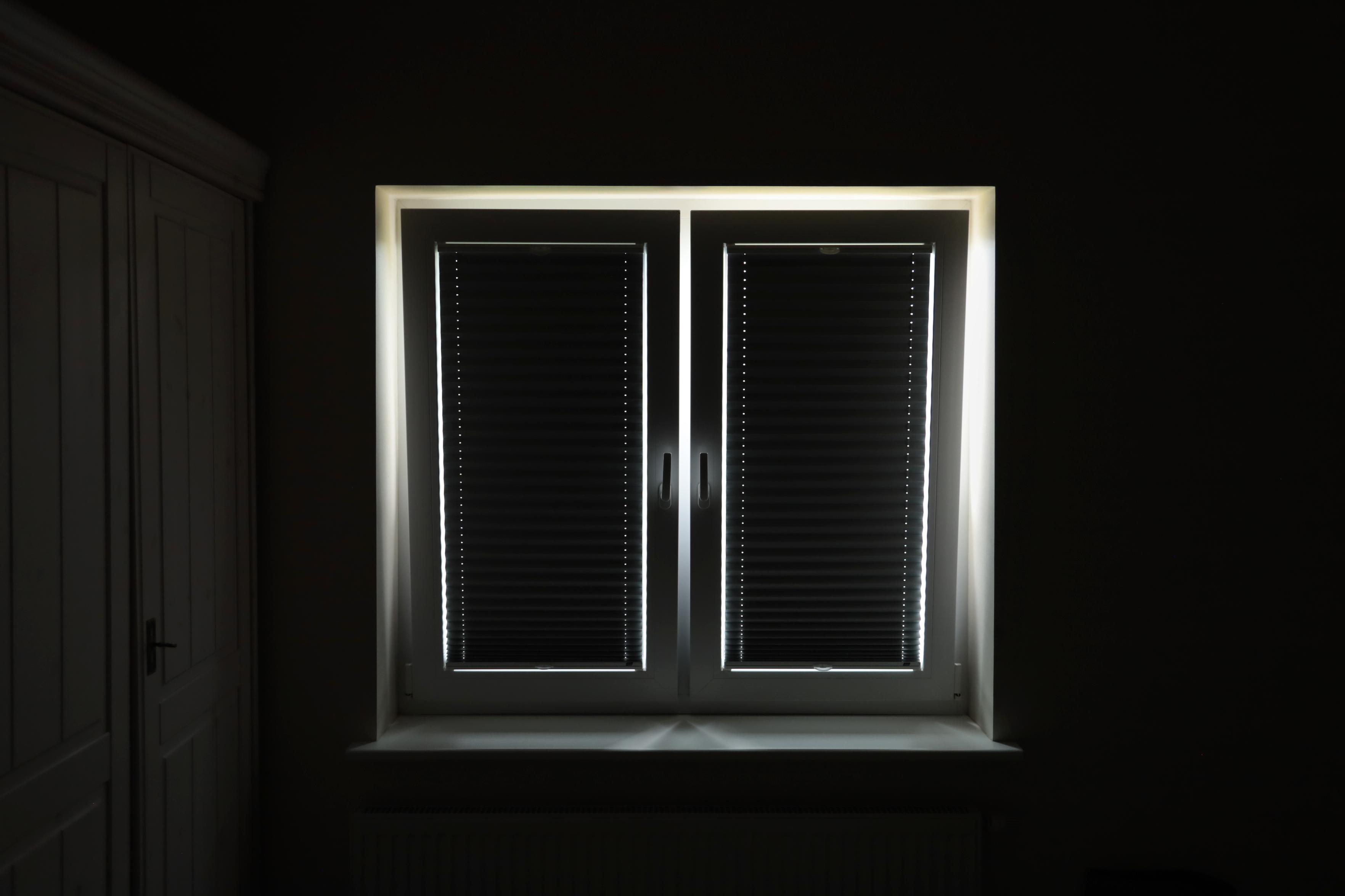 Schwarzweiss-Foto eines Doppelfensters von innen. Die Verdunkelungs-Plissees sind über die ganze Glasfläche aufgezogen. Von außen scheint das Sonnenlicht durch die umlaufenden Grenzflächen. Beide Fensterflächen sind in Kipp-Stellung. Auf der linken Seite neben dem Fenster sind Teile eines Schrankes schemenhaft zu erkennen.