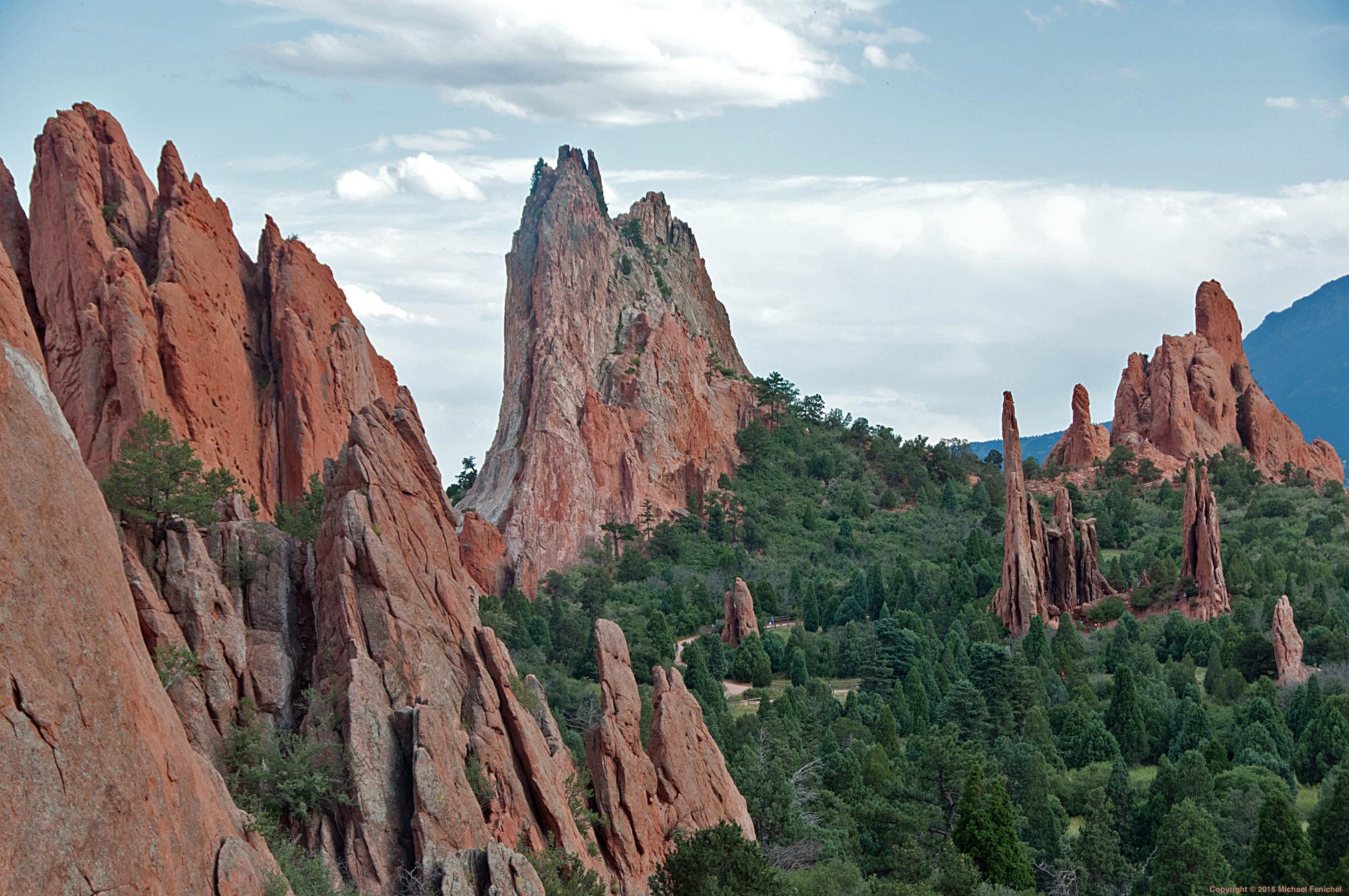 Photo by Fenichel:

Reddish (sandstone) rock formations in the Garden of the Gods

The tall center monolith is the Cathedral Spires, with Slender Spires to the right, and Three Graces far right.
 
"The massive, sloping red sandstone rock wall framing the left side of the view is part of the South Gateway Rock" - Alamy

Along the center and bottom are trees, and above is a bluish sky and white clouds.