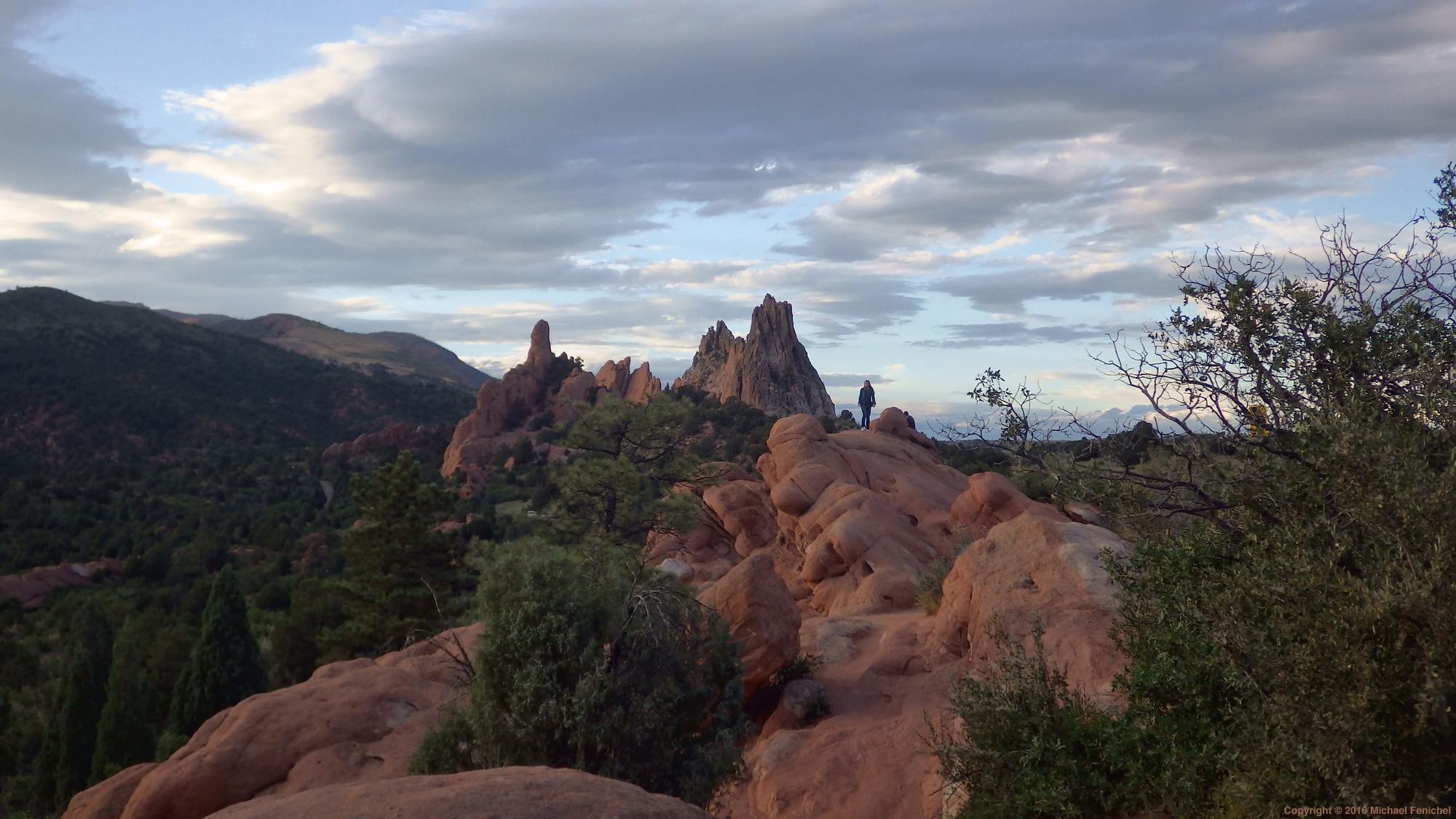 Photo by Fenichel:
Image of red, roundish rock in the foreground and spires in the distant background, with a lone figure standing atop a formation (center).

Below are trees, above sky with many clouds.