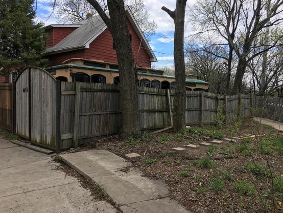 a photo of a house behind a wooden fence. behind a fence, two trolley cars can be seen peeking over the top. there is a gate at the front to let the trolleys out for a walk