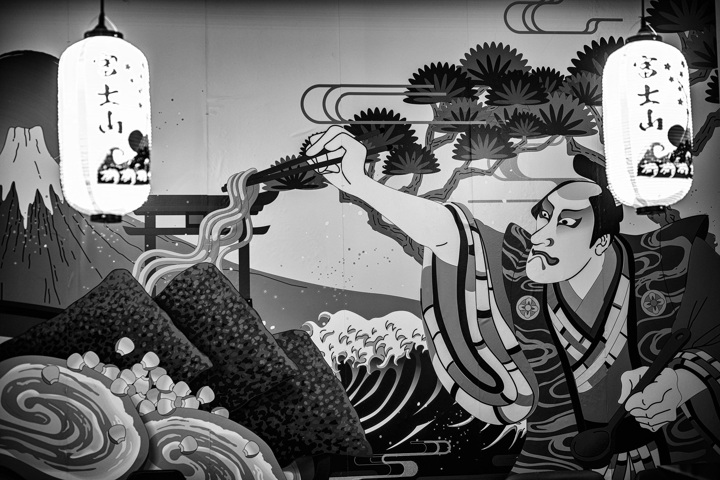Black and white photo in landscape format of a mural inside a ramen restaurant.

Two Japanese chouchin (提灯), or paper lanterns, are hanging from the ceiling in front of the mural in the top left and right of the photo. 富士山 (Mt. Fuji//Fujisan) is written in Japanese vertically on the left of each lantern with an ocean wave and part of the mountain drawn on the right.

The mural has a chain of matsu (松) pine tree brannches extending from the top left corner to the middle of the frame. On the top left of the mural, a rising sun behind 富士山. 

Under the mountain are ingredients from the restaurant's signature ramen dish: three sheets of nori, two pieces of char sui, and corn. Above the nori sheets three strands of ramen noodles are held above the dish with a pair of chopsticks/hashi (箸) by a kabuki yakusha/actor (歌舞伎役者) right arm extended, left hand holding a soup both ladle at waist height, and dressed in traditional kimono. He's intently concentrating on the ramen as if finishing its pres