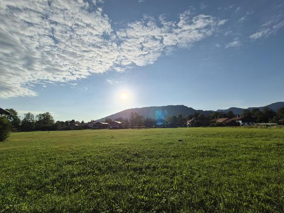 This image captures a wide, open meadow with vibrant green grass stretching toward a line of houses and trees in the background. The sky above is a brilliant blue, dotted with scattered white clouds. The sun is visible near the center of the image, partially obscured by a lens flare effect, casting a bright and warm light over the scene. Beyond the houses, rolling hills or mountains rise, adding depth and a sense of natural beauty to the landscape.

description generated with Mistral.ai and rev…