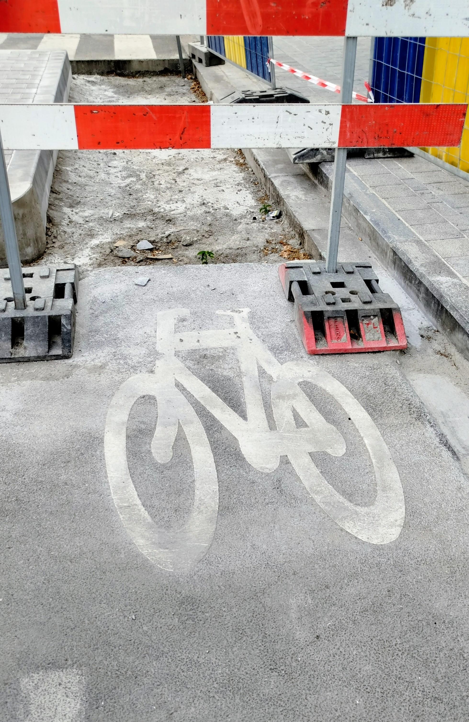 Dans la rue, travaux. Sur le sol le dessin de signalisation de vélo en blanc puis un panneau pour empêcher le passage et derrière la route sans bitume