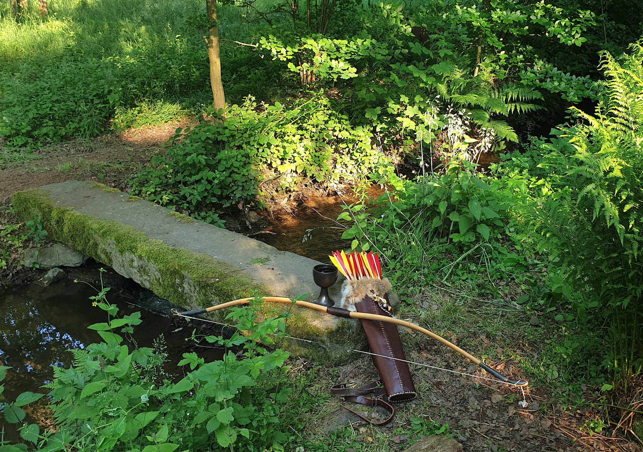 Tag. Bach. Viel Grün. Kleiner Steinsteg. Davor ein Langbogen aus Holz und ein Köcher mit Pfeilen mit geldroten Federn. Ein Tonkrug steht dabei.