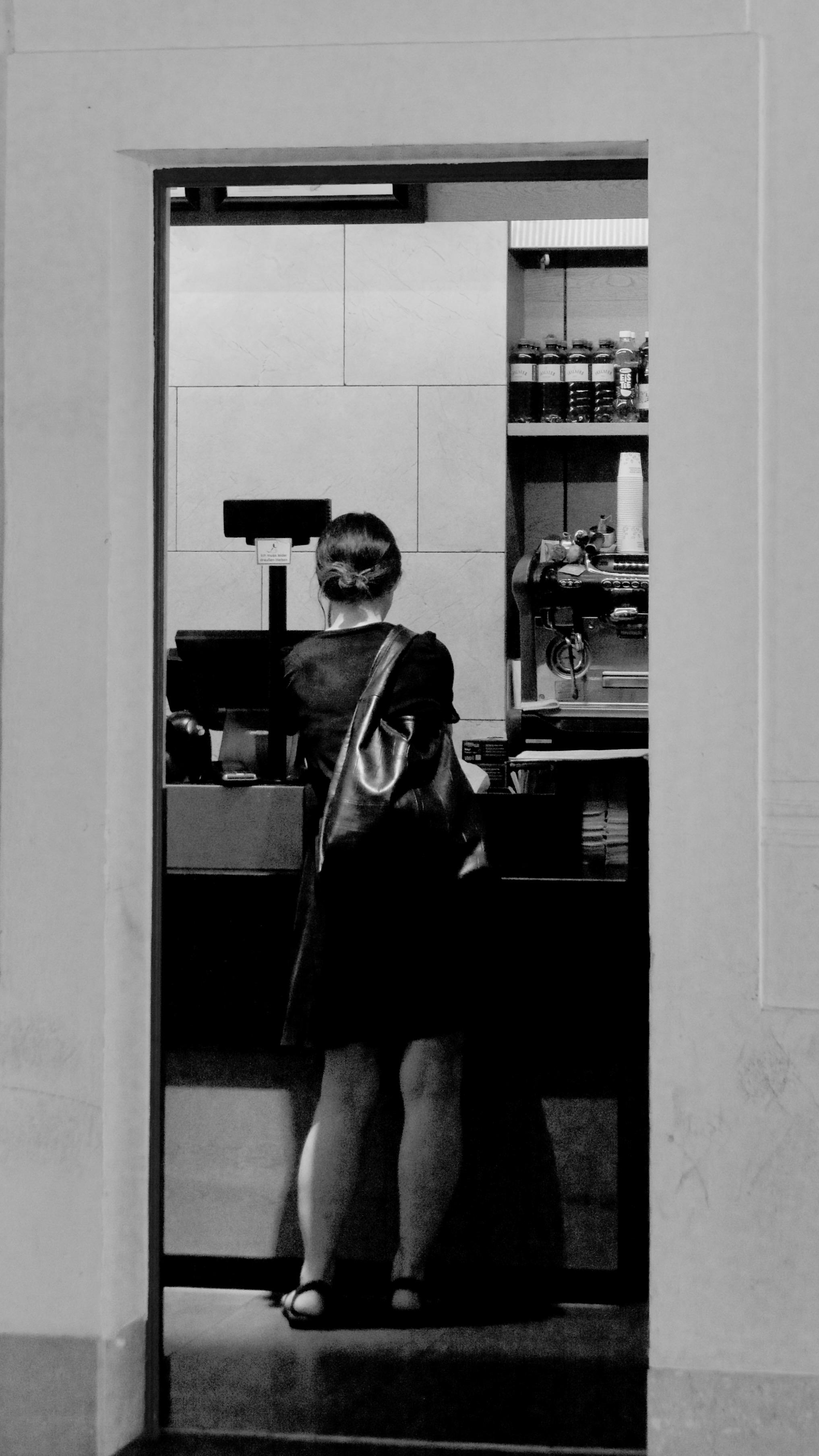 Picture of a young woman, buying a coffee in the morning. Photo is taken through the door (which is the frame of the shot) and black and white. The person is portrayed from behind, in front of a counter of a small bakery in a subway station in Vienna,