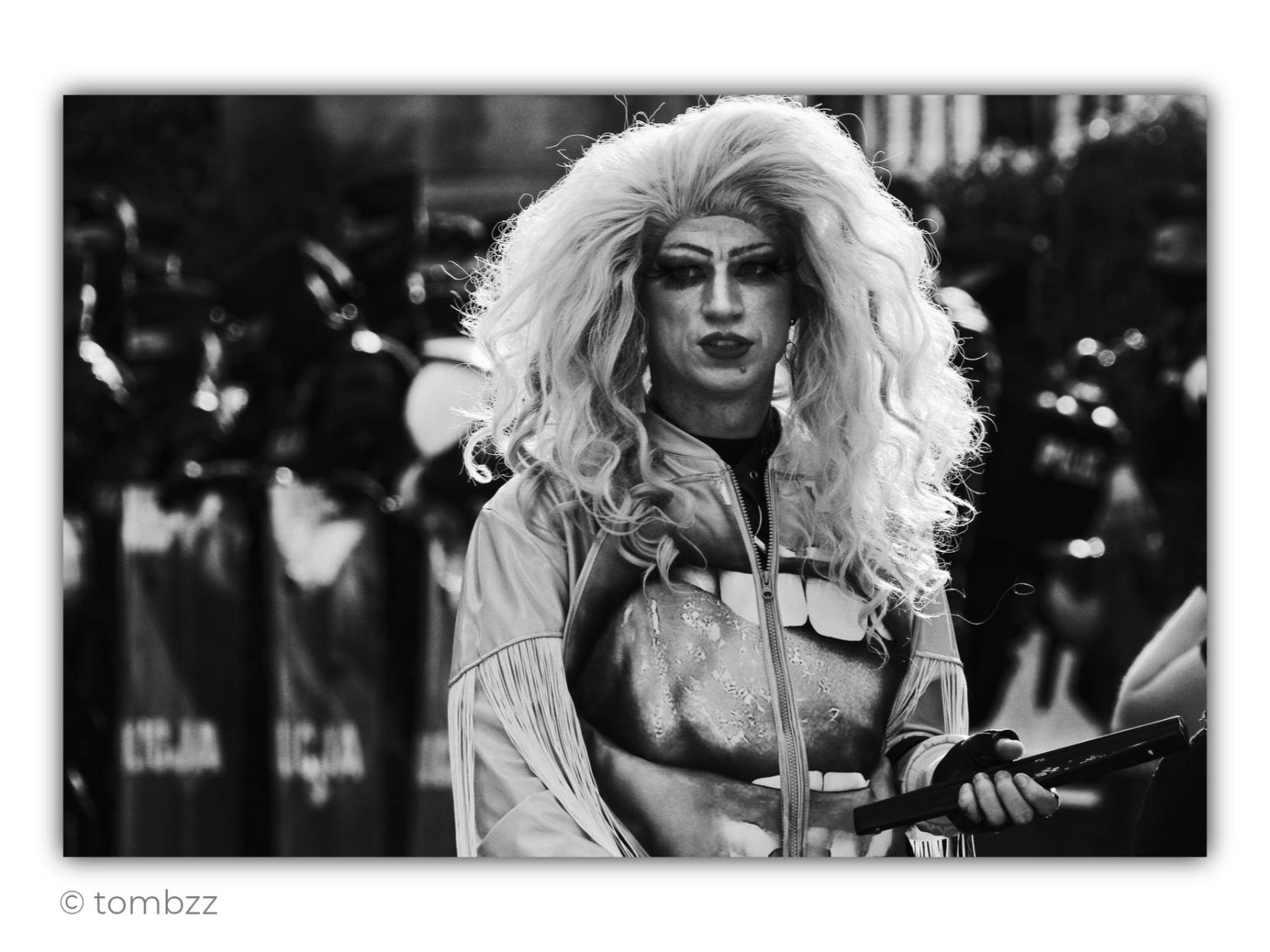 Black-and-white street photograph taken in Poland, just before the start of an LGBT+ Pride March. At the center of the frame stands a solitary person with a striking, theatrical appearance: a very light, voluminous wig, expressive eye and lip makeup, and a shimmering outfit with fringed details. They look directly into the camera, calm and unsmiling. In one hand, they hold a dark object resembling a fan.
Behind them, outside the depth of field, stretches a line of police officers in protective gear. Their dark silhouettes, helmets and shields form a heavy, almost uniform background for the brightly lit figure in the foreground.
The photograph was taken during a period of right-wing populist government in Poland, when LGBT+ people were subjected to intense political hostility and public rhetoric that opposed “people” to the so-called “LGBT ideology.” It was also a time of real aggression toward people associated with the rainbow and Pride marches. In this context, the solitary figure st
