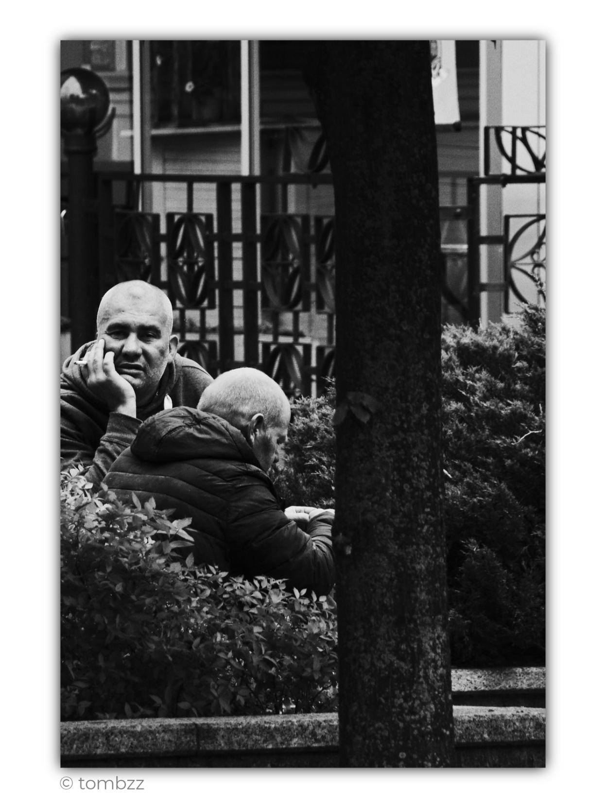 Black-and-white street photograph taken in a park. Two older men are sitting on a bench, partly hidden among low shrubs. The man on the left, with a shaved head and short stubble, rests his face on one hand while holding a cigarette between his fingers. He looks ahead with slightly narrowed eyes, as if the conversation has paused for a moment and he is lost in thought. The other man, noticeably older, sits turned sideways with his head bowed. He is wearing a quilted hooded jacket, and his quiet, focused posture suggests that he is listening or reflecting.
A thick tree trunk cuts through the foreground, partly obscuring the two men and dividing the frame into separate areas. Dense shrubs surround the bench, while a metal fence and fragments of buildings can be seen in the background. The composition feels like a moment observed discreetly from a distance, an ordinary fragment of everyday life. Two people, a conversation, a gesture, the silence between words, and a park that briefly beco