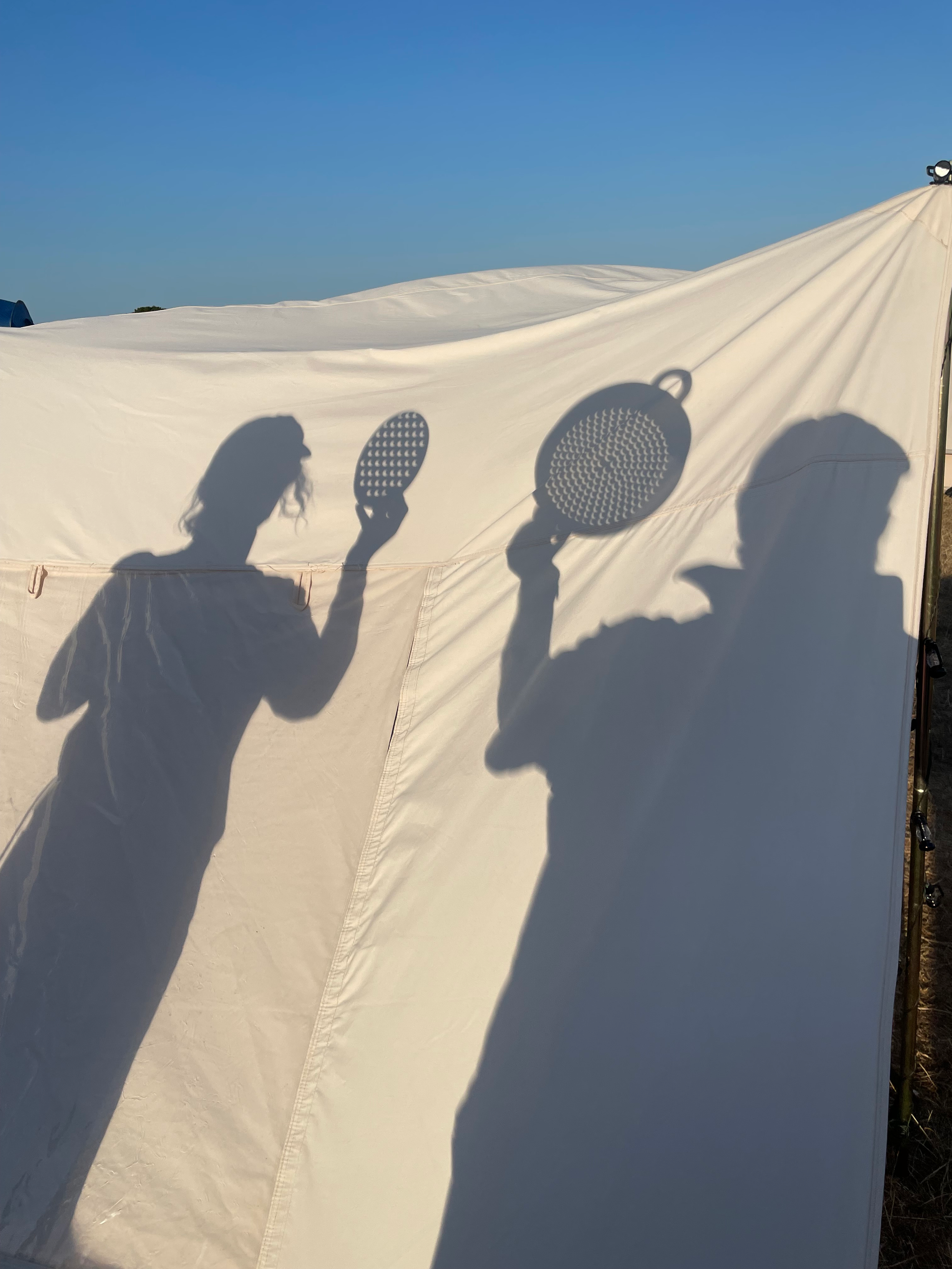Shadows of two people on cream coloured canvas. One shadow figure holds up an oval cheesegrater, the other holds up a round colander. The holes in each are clearly visible as a pattern of crescent shaped dots, each being a roughly focussed image of the sun part way through the eclipse. So there are hundreds of eclipse images. The canvas takes up most of the picture, and is topped by a blue sky.