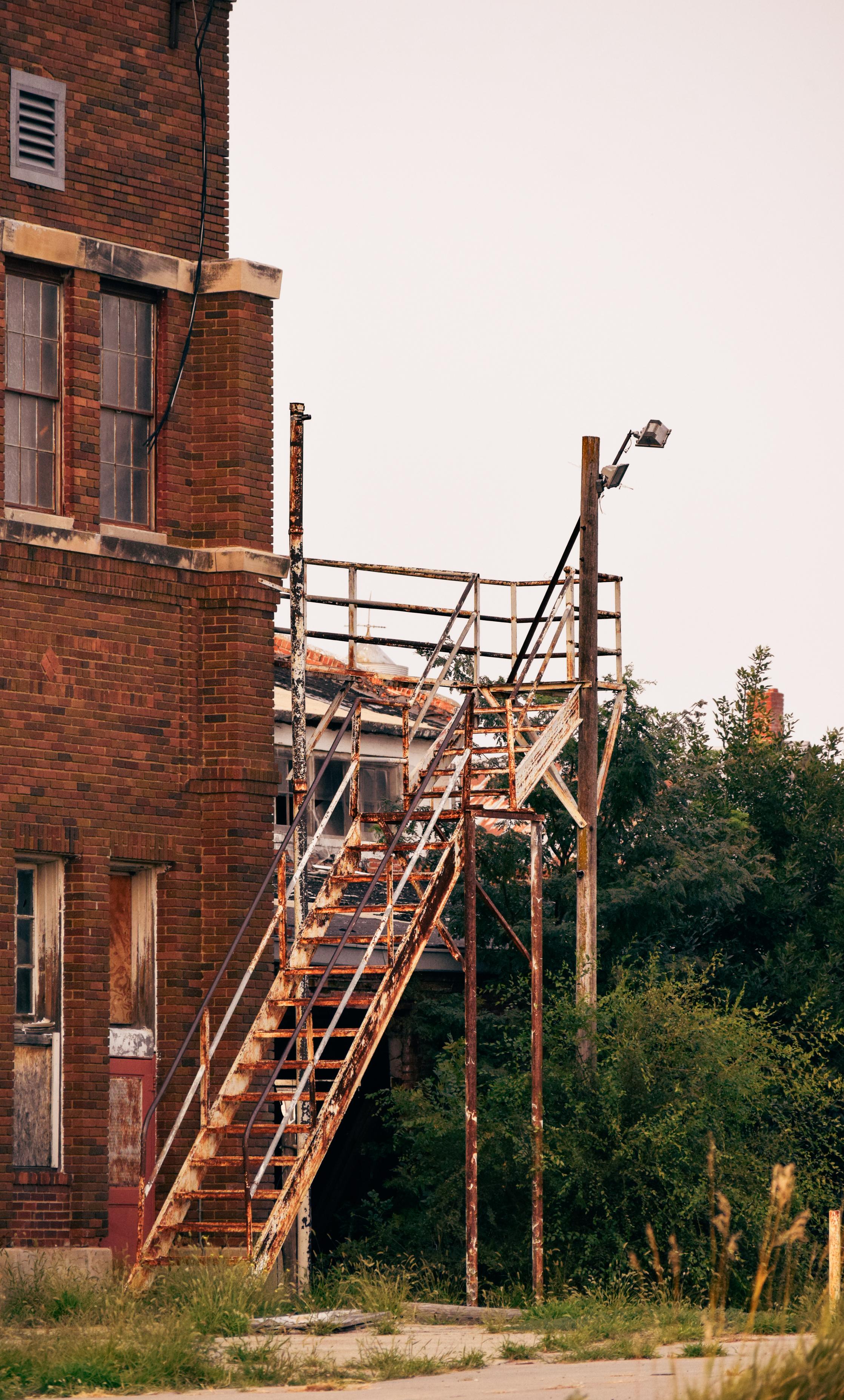 An aging red-brick industrial building with rusted metal fire stairs and railings, set beside overgrown greenery under a pale sky.