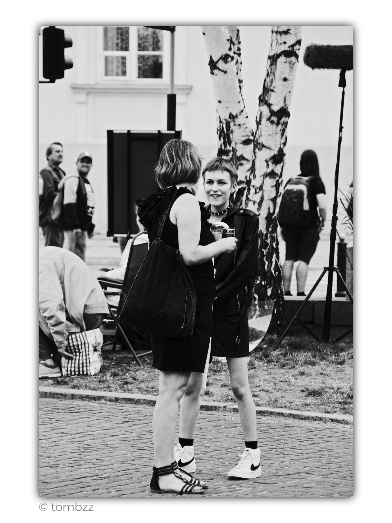 Black-and-white street photograph taken on August 1, the anniversary of the outbreak of the Warsaw Uprising. In the foreground, two young women stand absorbed in conversation. One, seen from the back and slightly from the side, has short light-colored hair, a black dress, and a large bag over her shoulder. The other stands facing her. She has a very short haircut, a dark jacket, a plaid skirt, light-colored sneakers, and a necklace with metallic details. She looks at her companion with a slight smile. Both women are holding memorial candles.
In the background, an ordinary city scene unfolds. People pass by, someone is sitting near a bench, and behind the figures there is the light facade of a building and two birch trunks cutting through the frame with strong, graphic patterns. On the right stands a tall microphone boom with a furry windscreen, used for recording sound outdoors.
Just outside the frame is the Warsaw Uprising Monument. The candles in the women’s hands give this seemingly