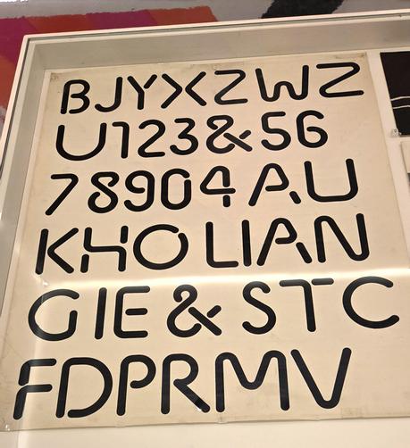 Photograph of a font specimen poster, shown in a display case. Some Photoshop has been applied to remove nasty reflections from the glass display case cover.

The poster reads:
BJYXZWZ
U123&56
78904AU
KHO LIAN
G IE & STC
FDPRMV