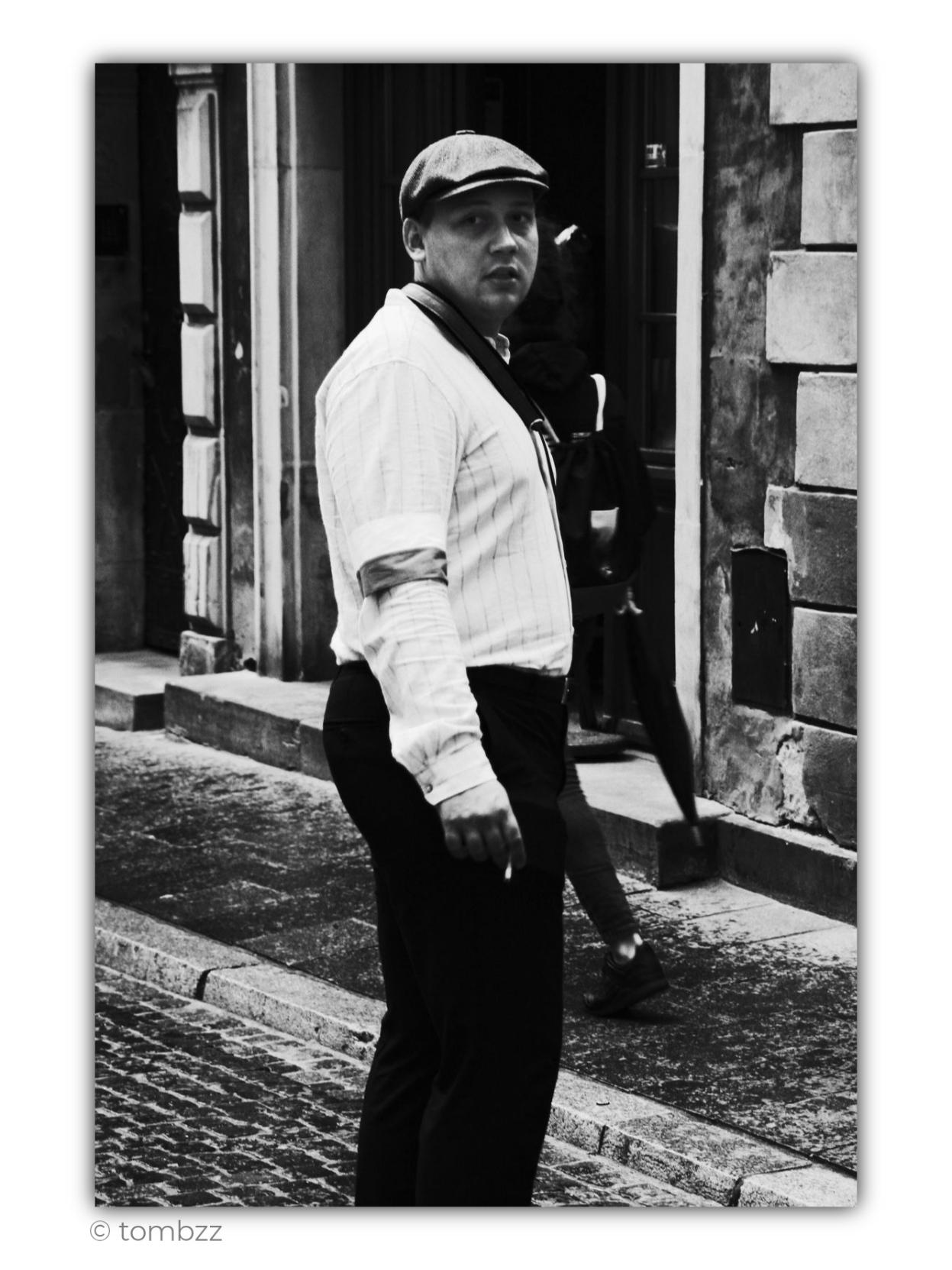 Black-and-white street photograph taken on August 1, the anniversary of the outbreak of the Warsaw Uprising. In the foreground stands a young man wearing a stylized outfit referring to the clothing of the insurgents of 1944. He wears a light shirt with rolled-up sleeves, dark trousers, and a characteristic flat cap with a short brim. A white-and-red armband is visible on his upper arm, one of the most recognizable symbols of the Warsaw insurgents. A dark strap, probably from a bag or part of his equipment, runs diagonally across his chest.
The man is captured in motion, as if he has just stopped and turned toward the photographer. He looks directly into the lens with a serious, slightly surprised expression. His figure stands out strongly against the dark entrance of an old tenement building. Behind him are tall wooden doors set deep into the façade, partly hidden in shadow. Another person is emerging from the doorway, visible only in fragments, adding a sense of movement and everyday 