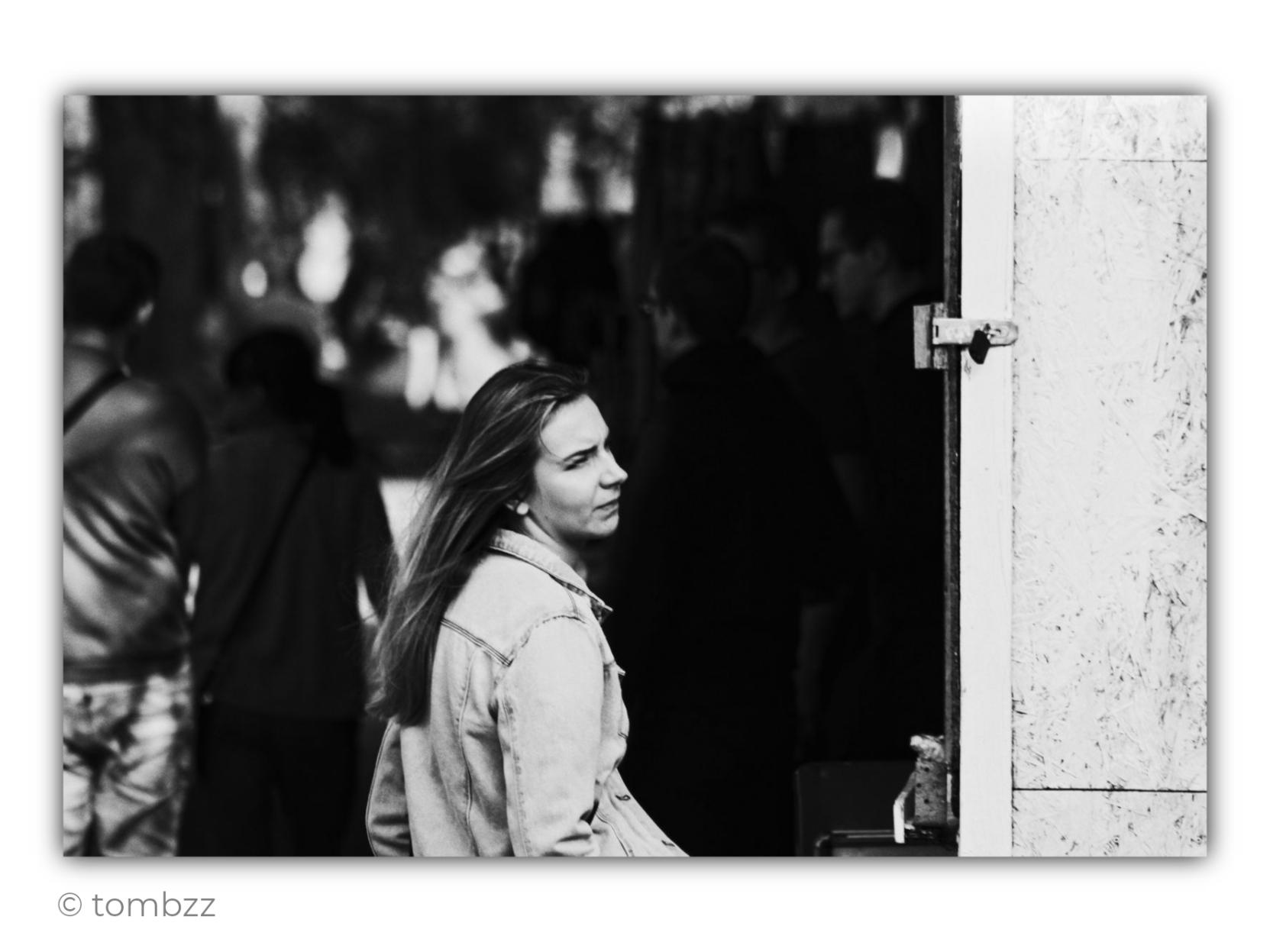 Black-and-white street photograph. In the foreground, a young woman stands brightly illuminated by sunlight, clearly separated from the shadowy surroundings. She has long, loose hair falling naturally over her shoulders and back. She is wearing a light denim jacket. Her face is seen in profile, turned toward the right side of the frame. Her gaze appears focused, slightly wary or curious, as if she is watching something outside the photograph.
Behind her stretches a deep, dark space filled with people. The figures closest to the camera almost merge with the shadows, while farther back there are patches of sunlight and brighter silhouettes. The background is soft and out of focus, turning the anonymous crowd into an impression of movement and everyday street life. On the right side of the frame is a bright section of a bar or street stall wall, with a visible metal hinge. The strong contrast between white, deep black and intermediate shades of grey creates a striking composition in which