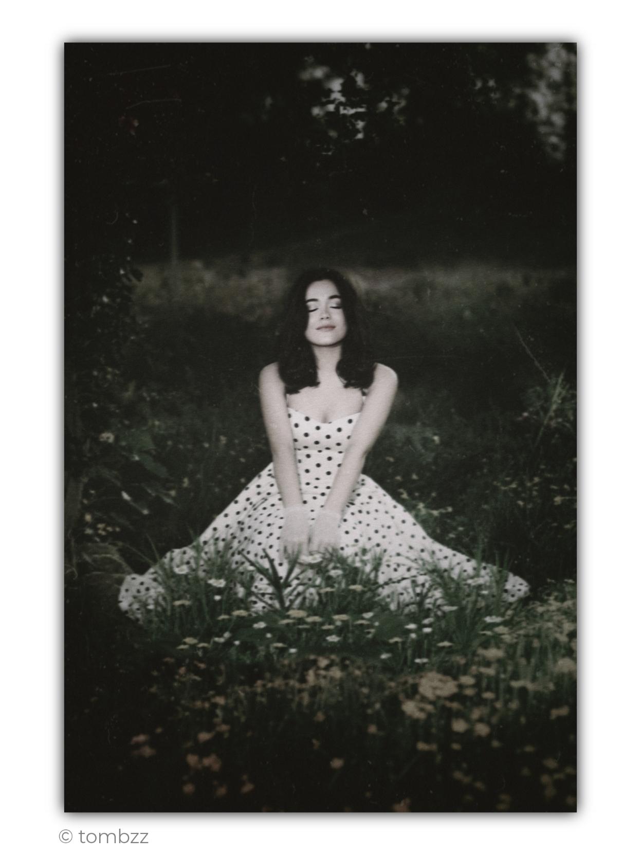 Analog portrait of my wife, Elisa, made among tall, freely growing vegetation. The colour palette is deliberately subdued, almost monochromatic, with cool greens fading into greys and muted creamy tones. The colours evoke old, slightly faded photographic prints and give the image a distinctly vintage character.
Elisa is sitting among grasses and small wildflowers, with her dress spread widely around her. The light-coloured polka-dot dress, fitted at the top and widening toward the bottom, strongly recalls feminine fashion of the 1950s. Delicate lace gloves further emphasize the styling and reinforce the retro aesthetic. Her dark hair falls softly over her shoulders. Her arms are extended in front of her, with her hands resting close together among the plants. Elisa’s eyes are gently closed, and there is a faint, almost imperceptible smile on her face. She appears calm, as if for a moment she has withdrawn from the surrounding world.
Her bright figure and the pattern of the dress emerge