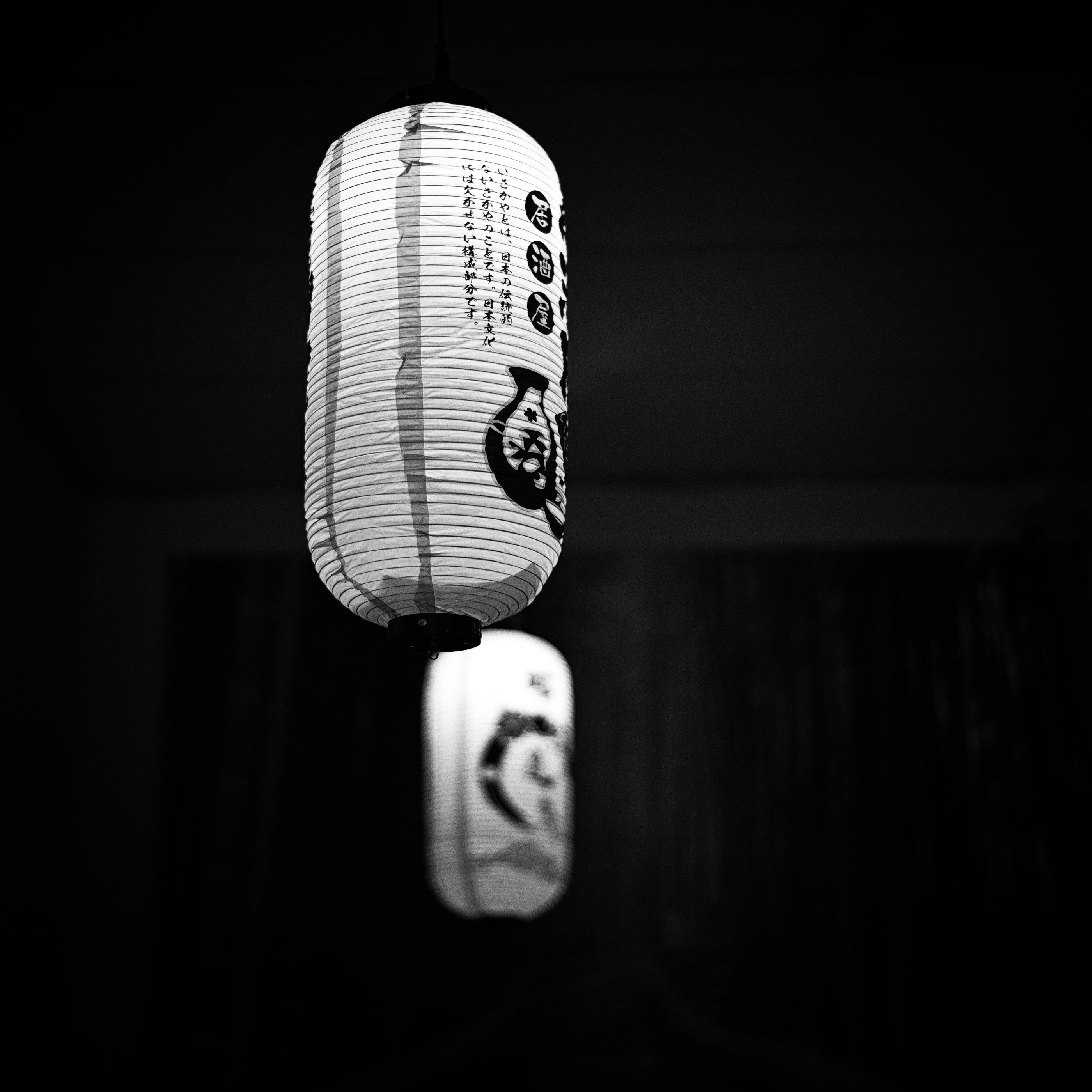 High contrast black and white photo in square format of two Japanese chouchin (提灯), or paper lanterns, hanging from the ceiling in a ramen shop.

The photo is metered for highlights and underexposed to give the feeling of a dimly lit room. A wide aperture was used to isolate the first lantern in the top half of the frame, which seems to be hovering in mid air, as the hanging ceiling bracket is barely visible. Two vertical painted lines on the left half, and small evenly spaced horizontal inner frames, run from the lantern's top to bottom. The right side has small vertical Japanese lettering in three columns, three more Japanese words circled in black, and a sake container to the right.

The second lantern, positioned below and slightly to the right of the first, is out of focus due to depth of field, but a dark colored crescent is visible in the center.

The far background has a barely visible painting with randomly shaped light colored vertical lines against a dark background, and two