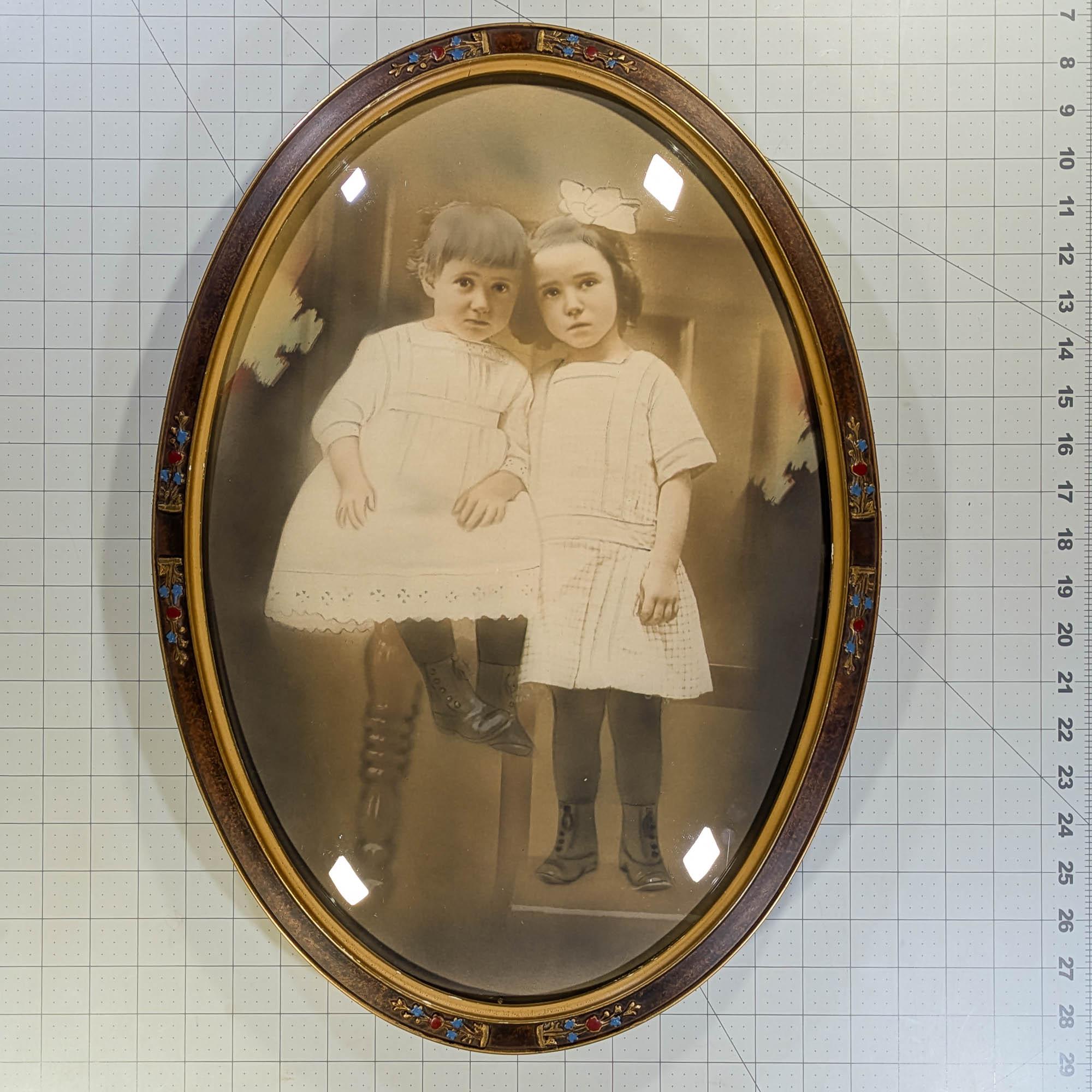 Photograph of two girls in a decorative antique bubble glass frame with red and blue flowers around the edge of the frame
Parts of the photo look to be enhanced by a crayon portrait artist