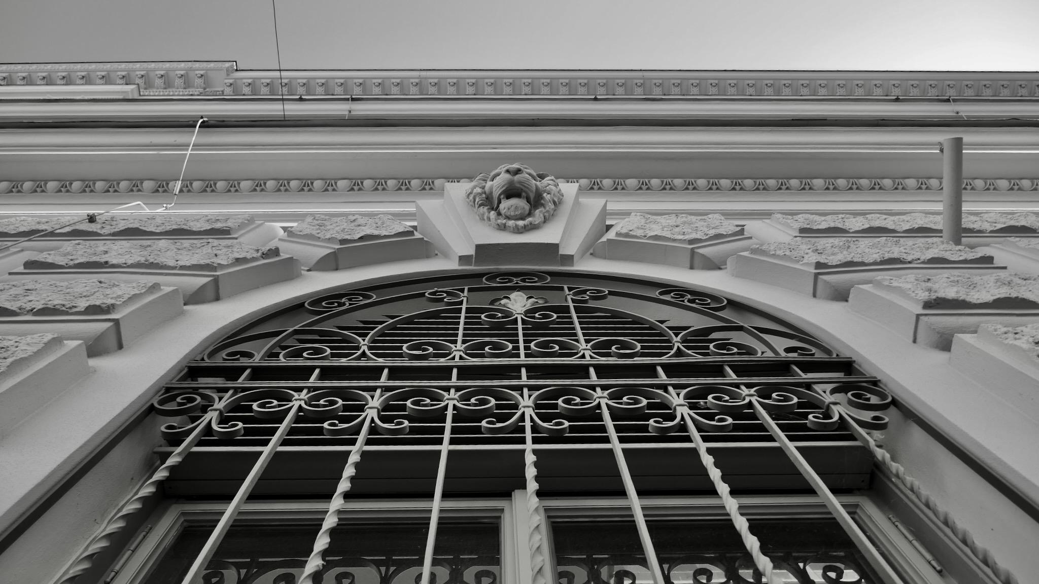 Picture of a window of an old building in Vienna. It is rounded to the top and there is a metal grid in front of it with ornaments. On the top is a stone lion head with an open mouth. Photo is taken from the ground up the facade. Picture is black and white.