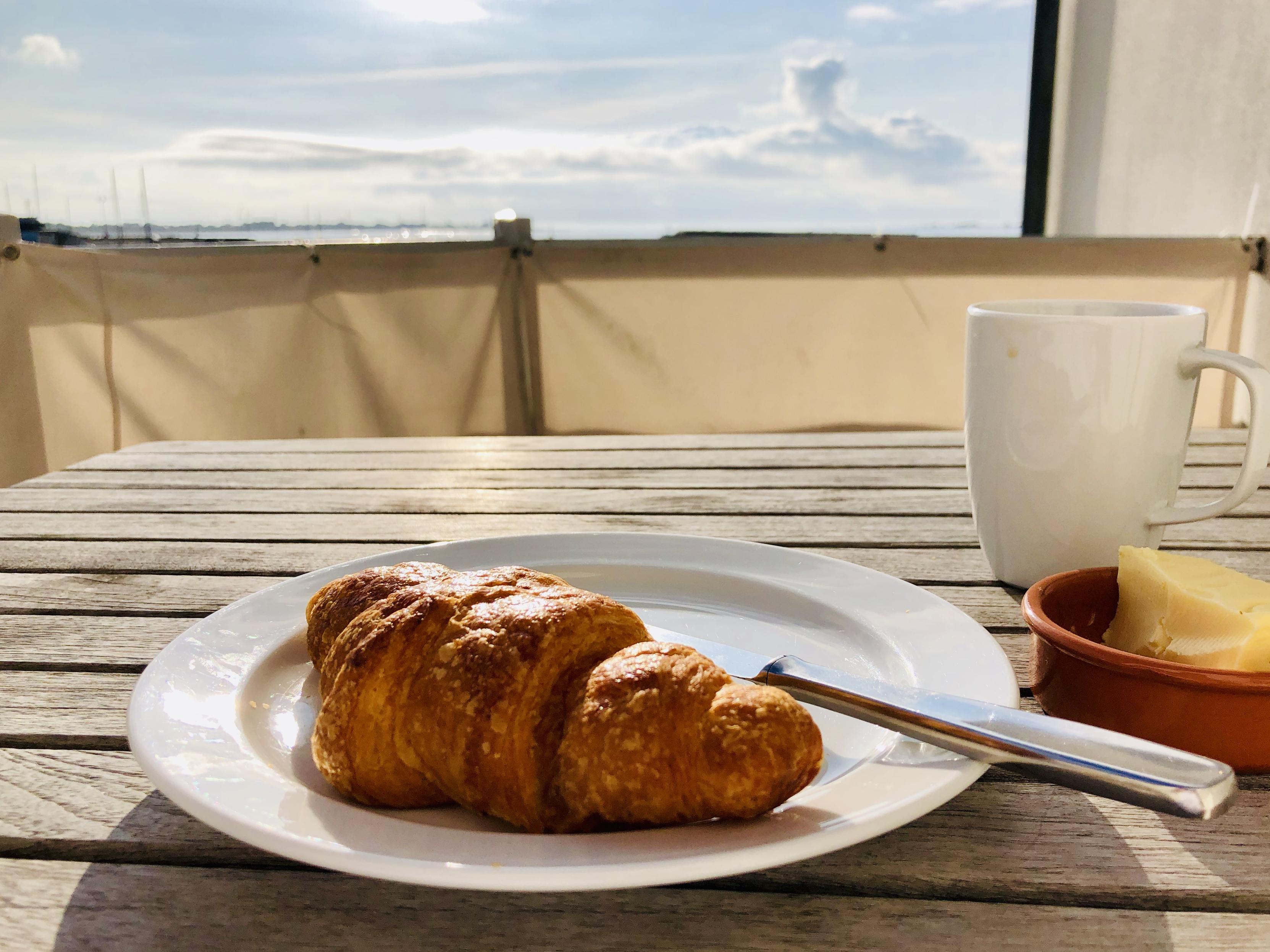 A croissant sits on a white plate on a wooden table. A white coffee mug and a dish of salty butter are beside it. In the background, there’s the Atlantic, and a partly cloudy sky.