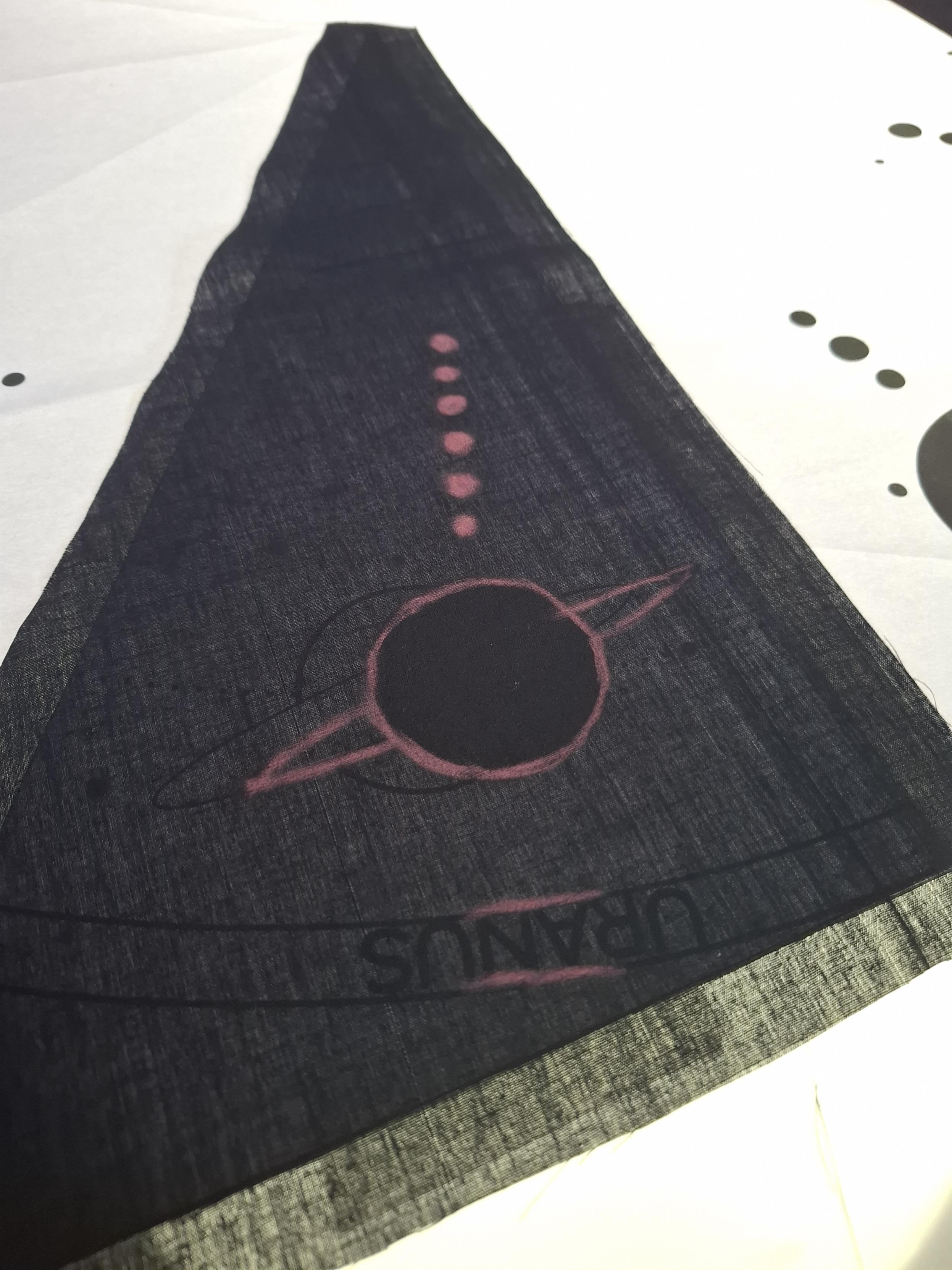 A piece of black fabric laid over two pieces of paper, one with the word uranus and a rough sketch outline, one with a black shape of uranus. There are markings on the black fabric in red chalk