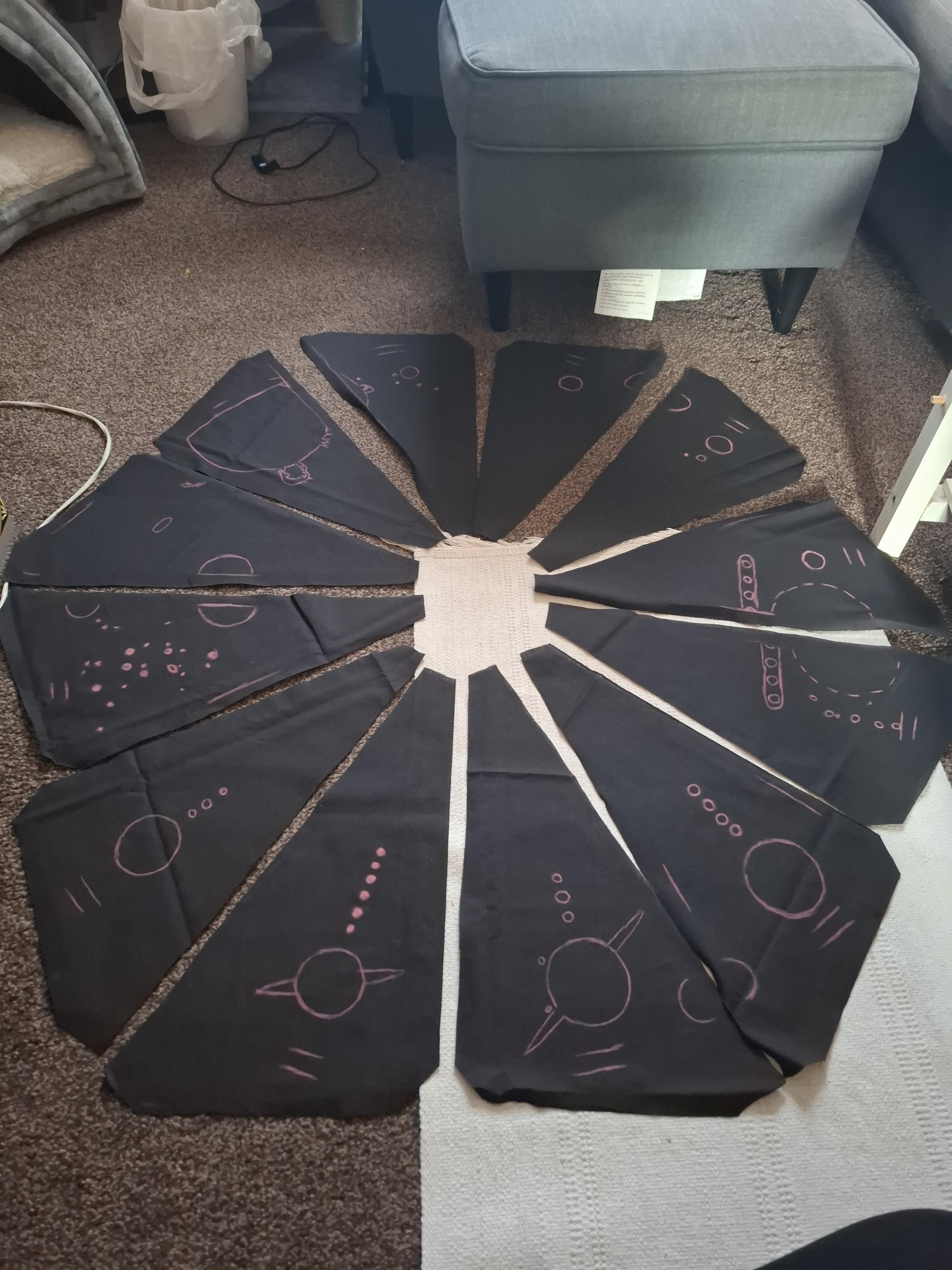 All of the sections of black cotton quilting fabric laid out on the floor in tildagon formation, with the different planets drawn on in red chall