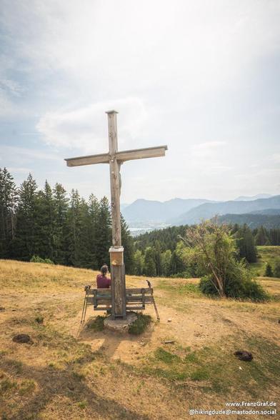 This image captures a wooden cross standing on a grassy hilltop with a panoramic view of a forested valley and distant mountains in the background. At the base of the cross, there is a wooden bench where a person is seated, facing away from the camera. The landscape is lush with evergreen trees, and the sky above is partly cloudy, creating a serene and open atmosphere.
description generated with Mistral.ai and reviewed with love