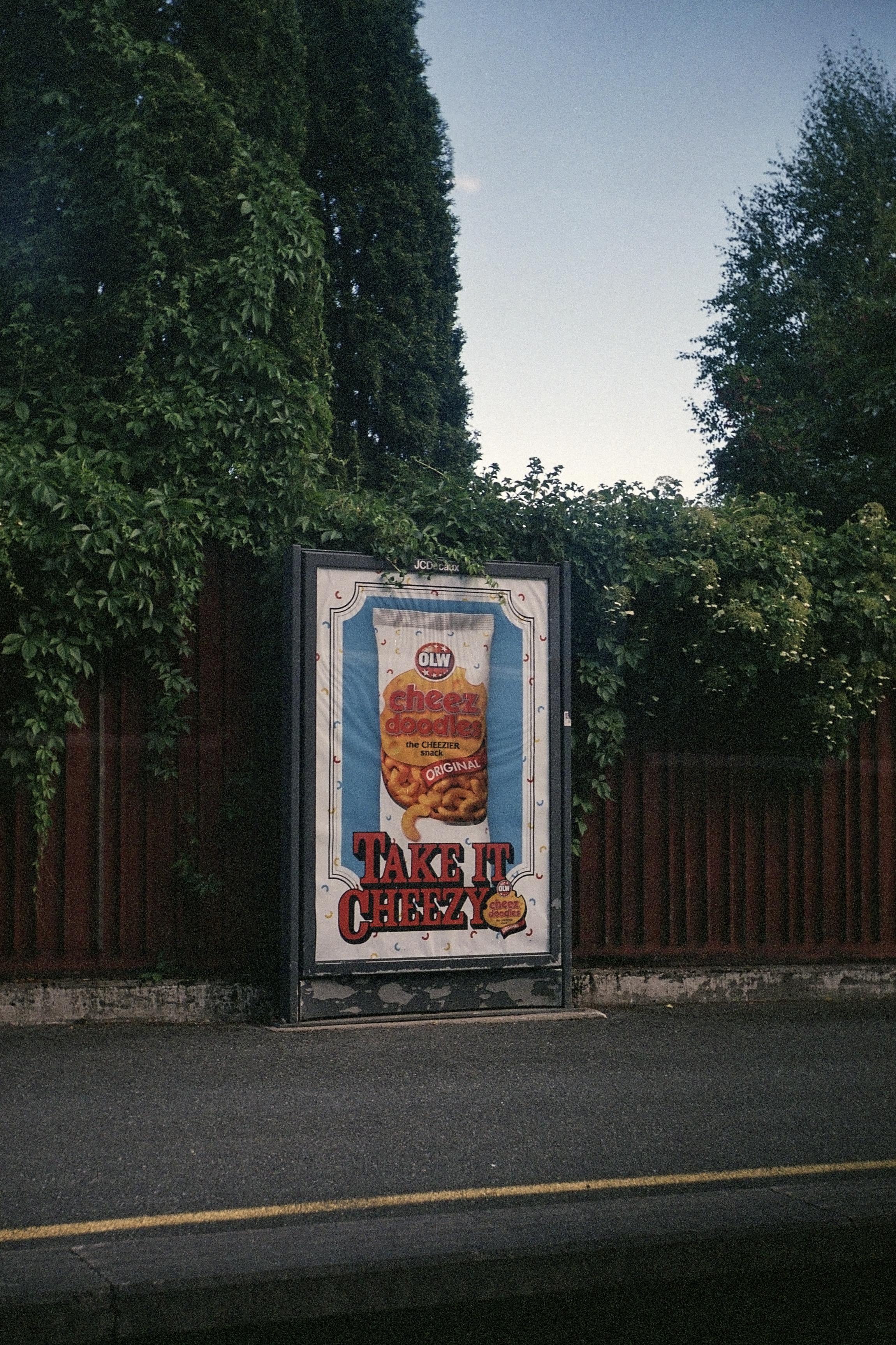 An advert for cheese doodles saying "Take It Cheesy" on an outdoor metro platform. Trees and a bit of sky in the background. Rainy summer weather.