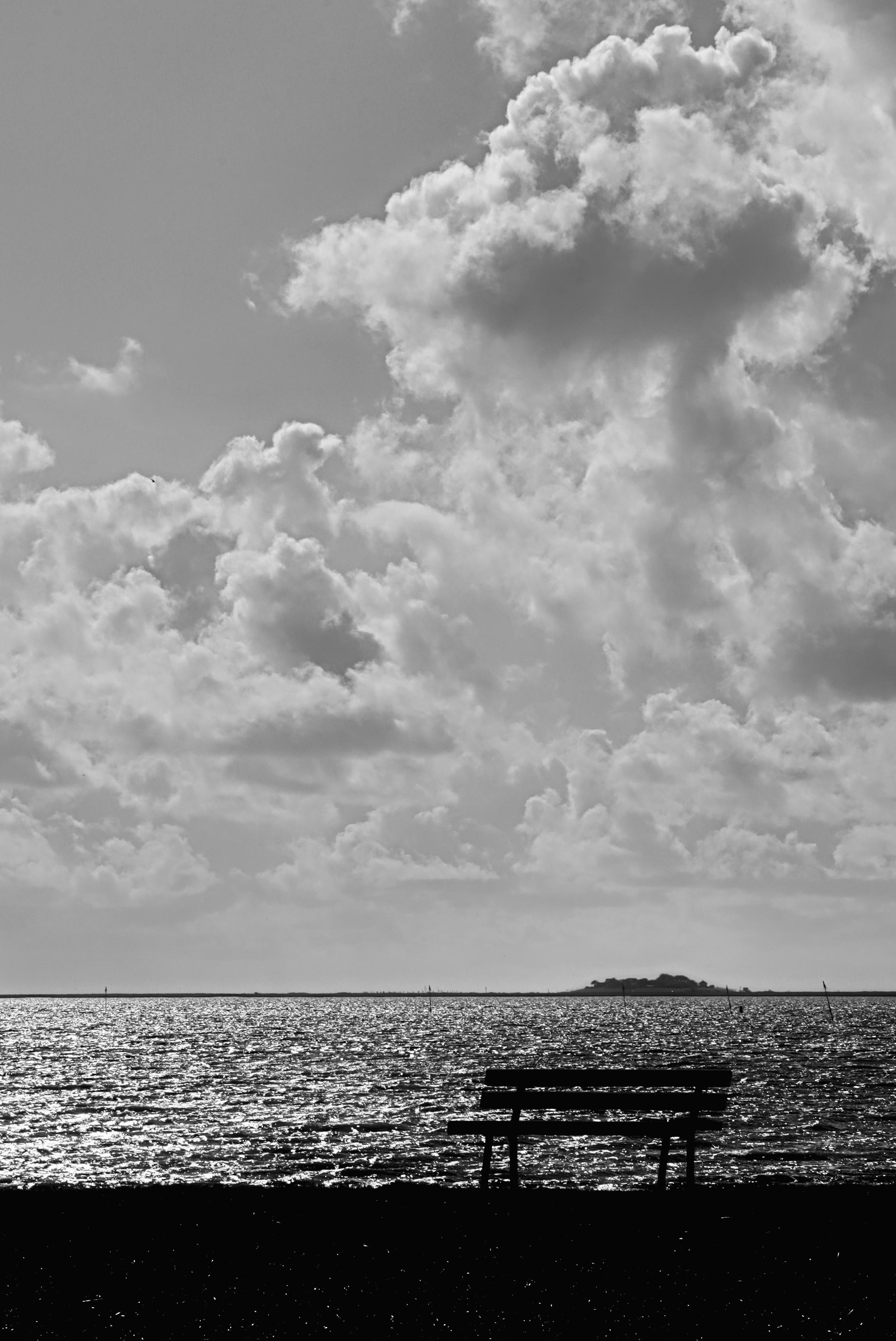 Schwarzweiss-Foto einer Sitzbank vor dem Wattenmeer (Nordsee). Über dem Horizont Wolkenstrukturen am Himmel. Am Horizont ein erhöhter Hügel (Warft) für Wohnhäuser auf einer flachen Insel (Hallig).