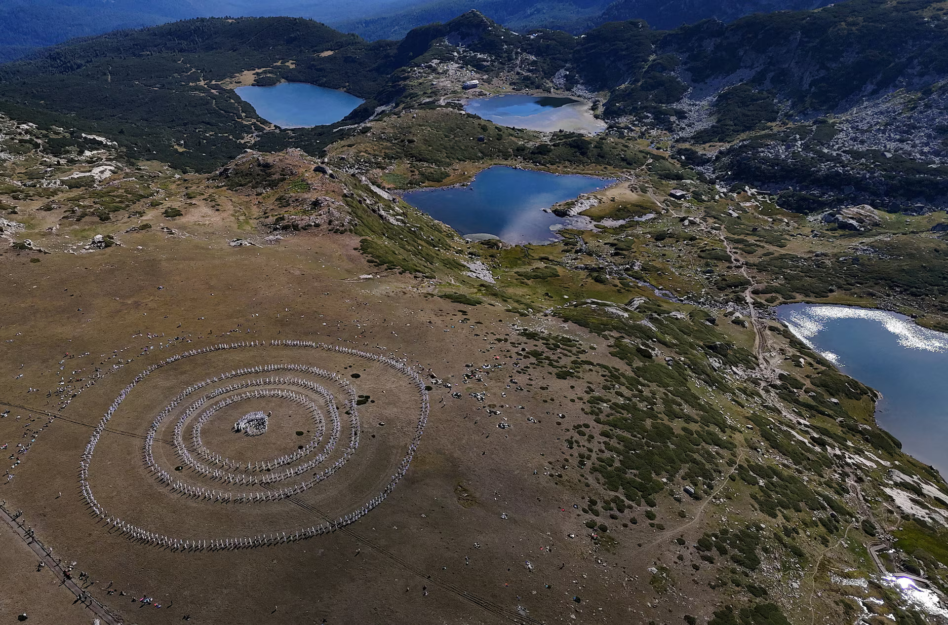 High-angle aerial view of a vast mountain landscape featuring several clear glacial lakes nestled among grassy, rocky terrain. On a wide, flat plateau in the foreground, hundreds of people dressed in white clothing stand in large, concentric circles forming a massive ritual or dance formation against the rugged natural scenery.